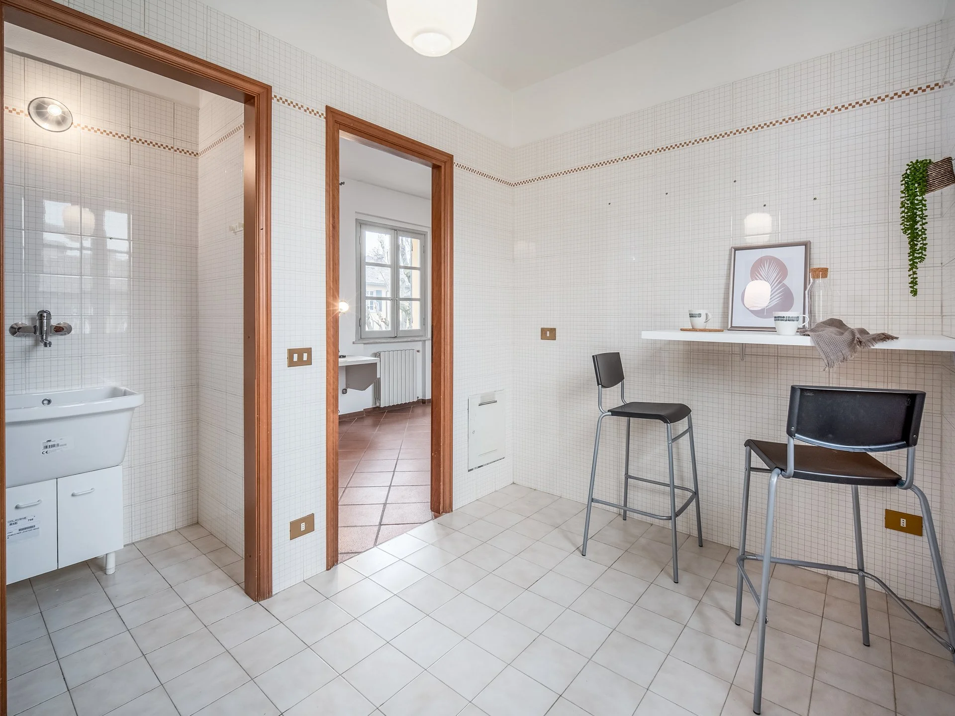 Interno di una stanza moderna con piastrelle bianche, due ingressi aperti verso altre stanze. A destra, un tavolo bianco con sgabelli, decorato con una pianta sospesa e una cornice con illustrazione. Ci sono accessori moderni sul tavolo.