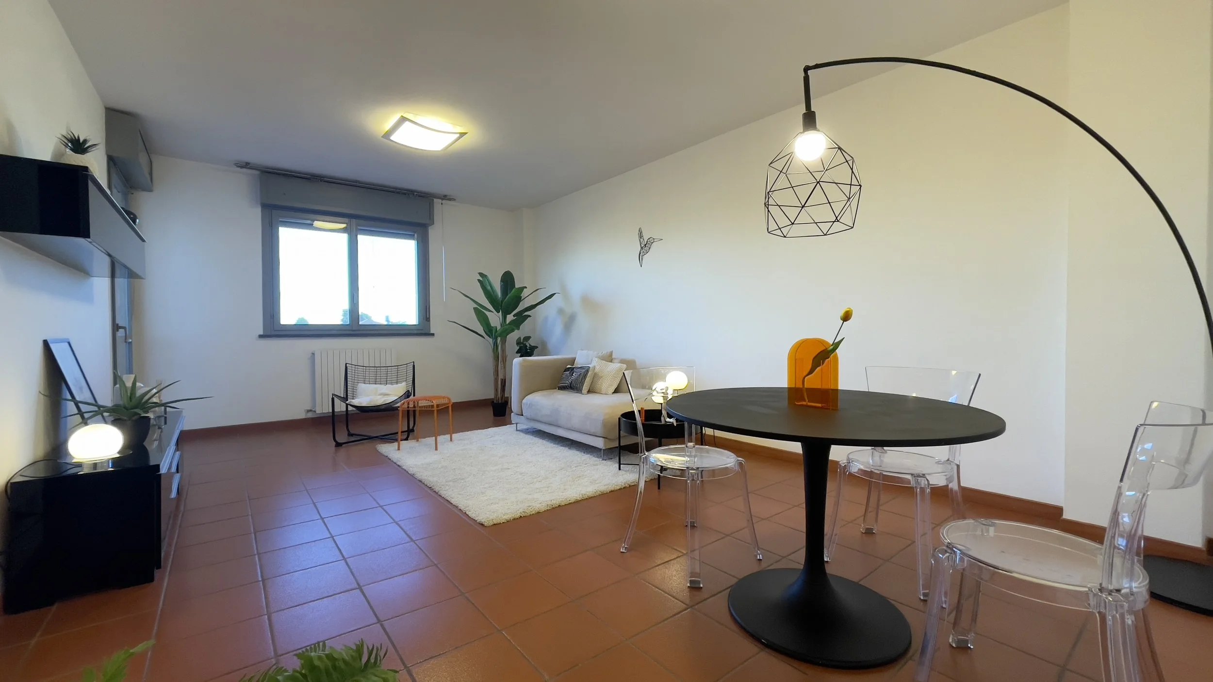 Soggiorno moderno con divano beige, tavolo nero rotondo con sedie trasparenti, pianta verde, lampada a sospensione e pavimento in cotto.