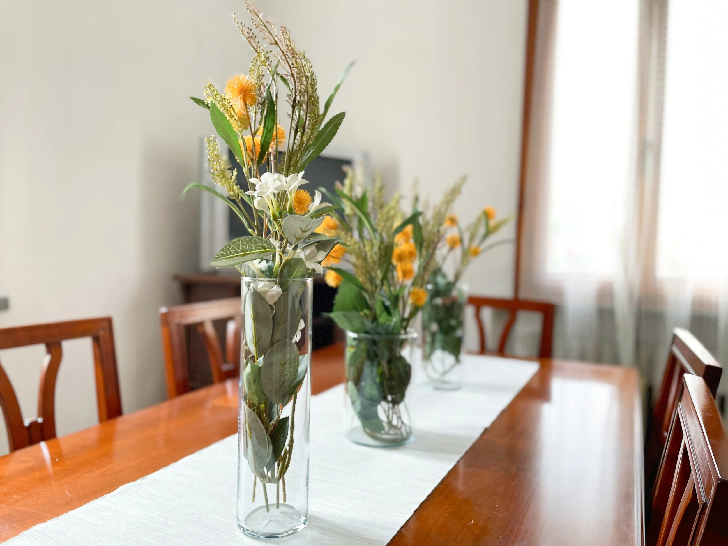 Tre vasi di vetro con fiori gialli e bianchi su un tavolo di legno in una sala da pranzo luminosa.