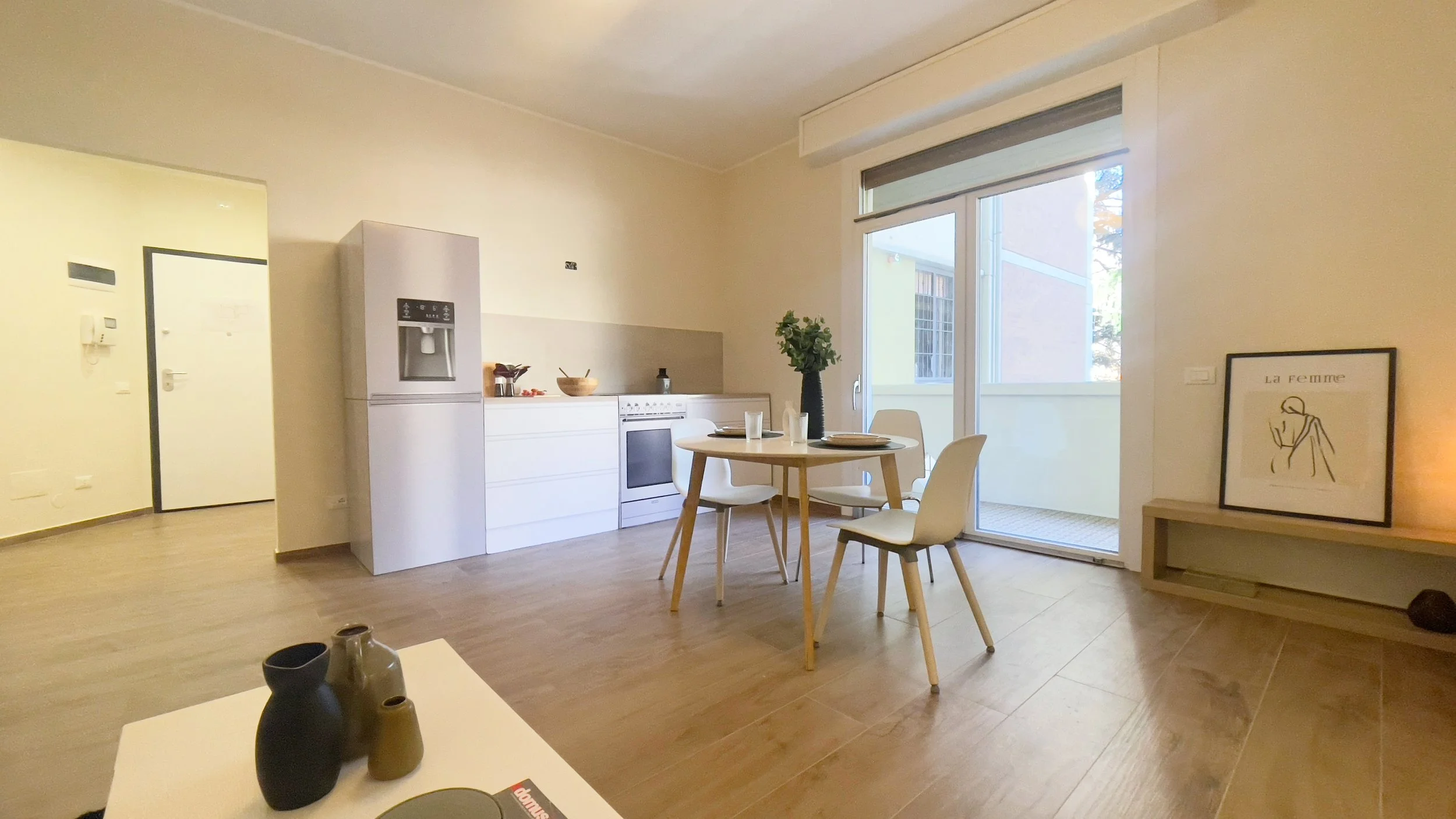 Cucina moderna con frigorifero, tavolo rotondo in legno chiaro e sedie bianche, finestra grande, pavimento in legno, e decorazioni minimaliste.