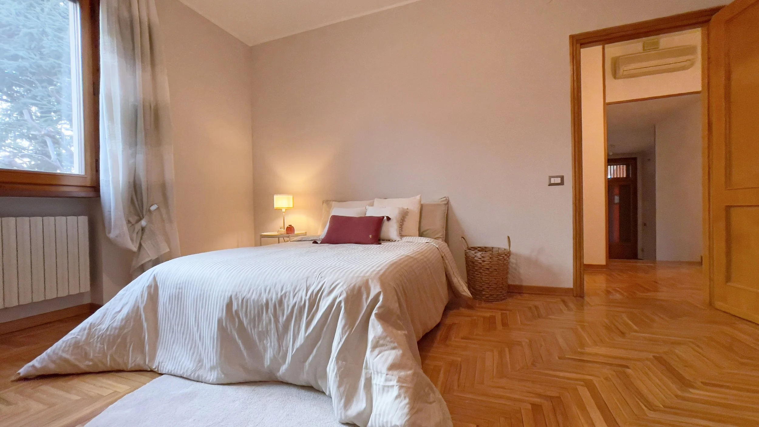 Camera da letto con letto matrimoniale, lampada da comodino accesa, pareti beige, pavimento in parquet e finestra con tenda bianca.