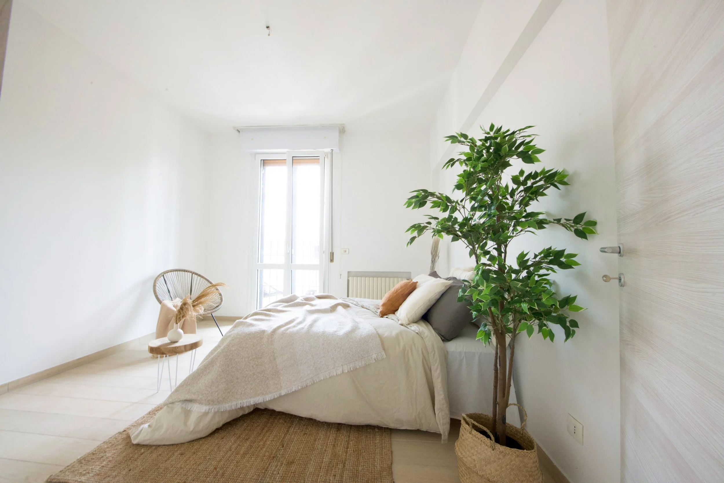 Camera da letto minimalista con un letto bianco, cuscini marroni, una pianta in vaso, una sedia di design e molta luce naturale proveniente dalla finestra.