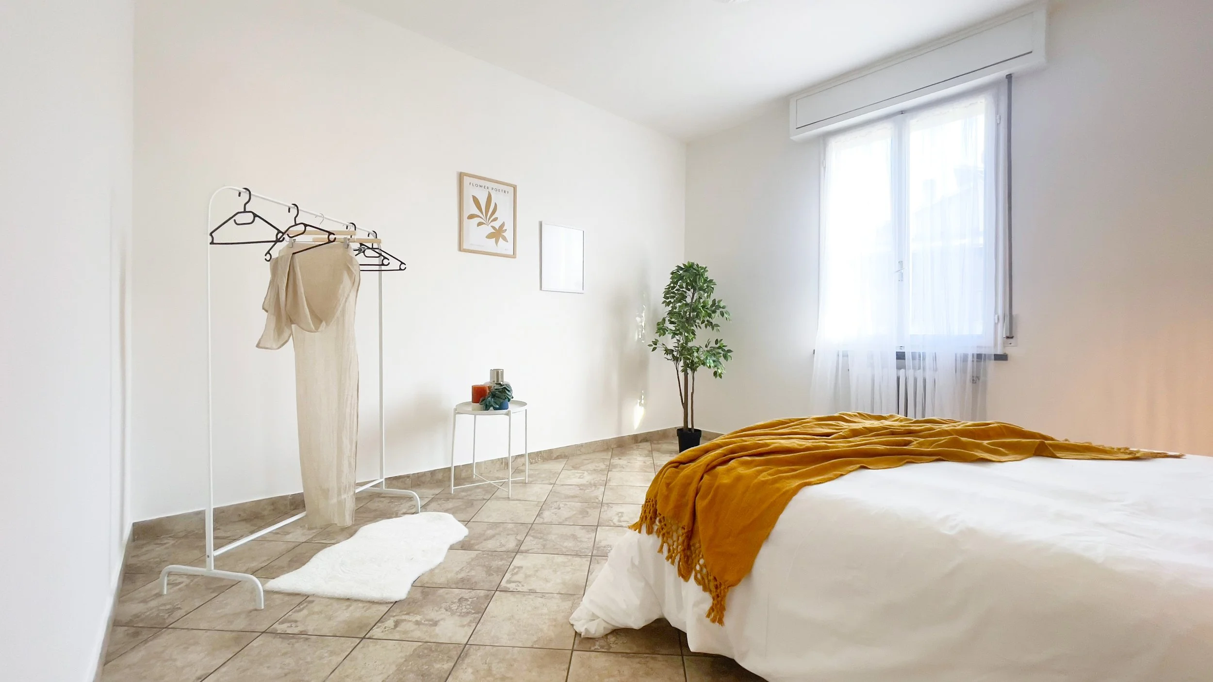 Camera da letto minimalista con letto, pianta, appendiabiti, finestra e pareti bianche.