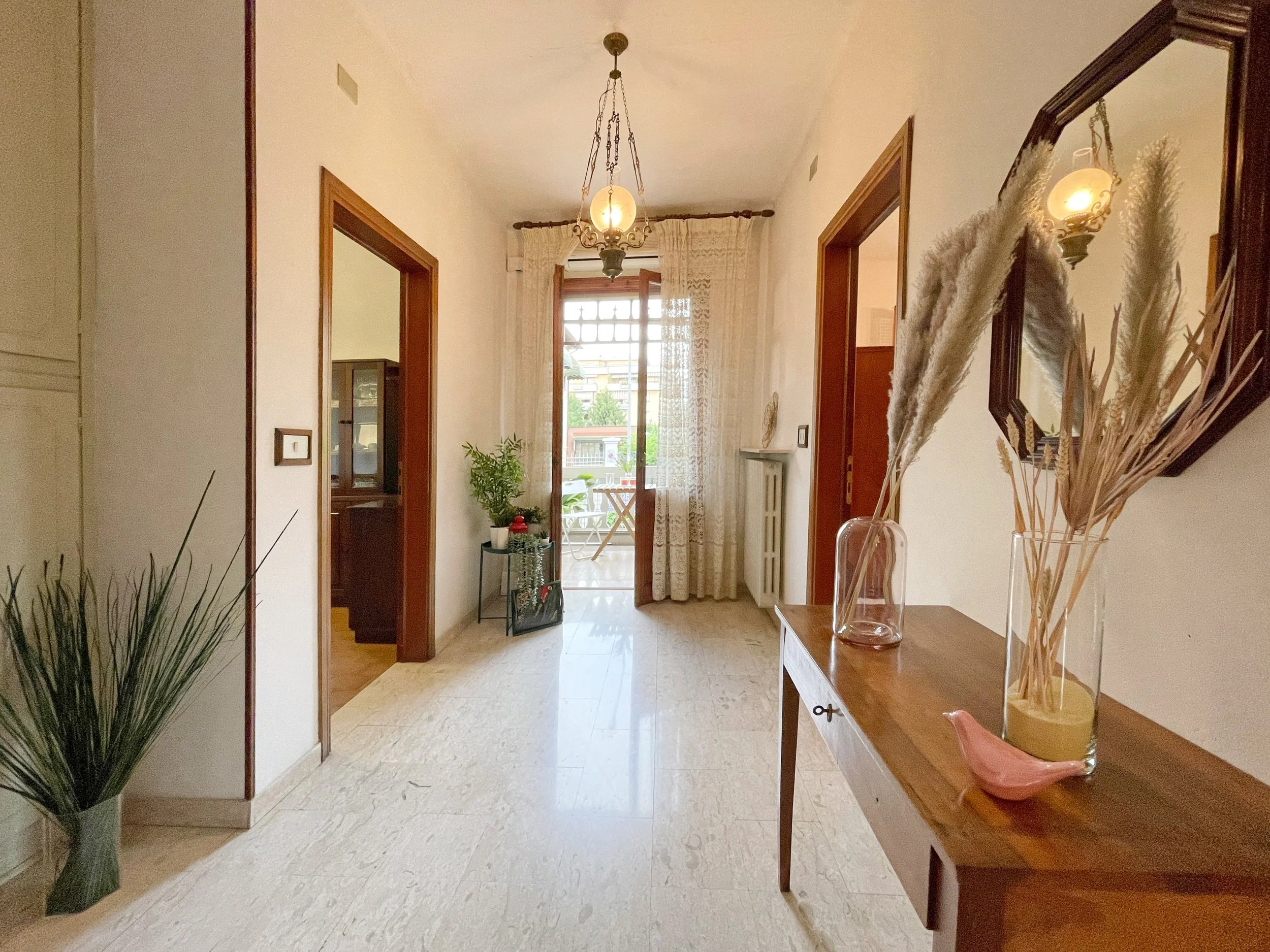 Interno di un ingresso di casa con pavimento in marmo chiaro. Ci sono un tavolino di legno con decorazioni, tra cui piume in vasi di vetro e un uccellino decorativo rosa. A sinistra, un vaso con piante verdi. In fondo, una porta finestra con tende ch