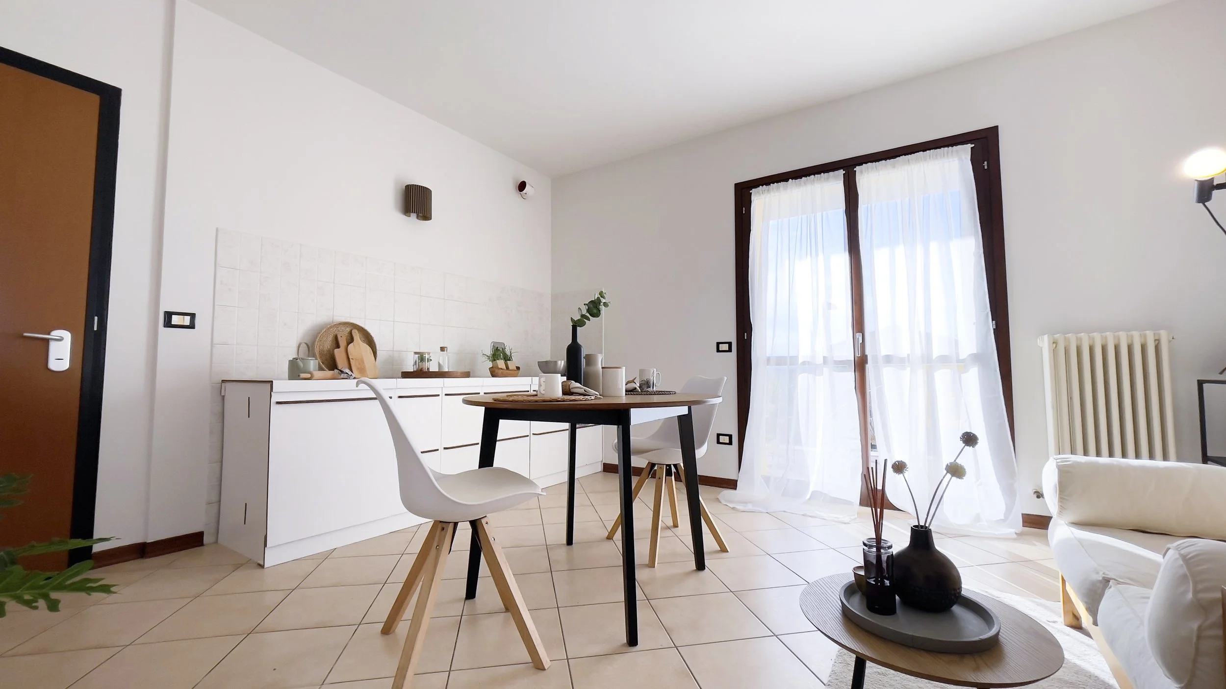 Sala da pranzo moderna con tavolo rotondo in legno, sedie bianche, finestra con tende bianche, radiatore e decorazioni minimaliste su un mobile contro il muro.