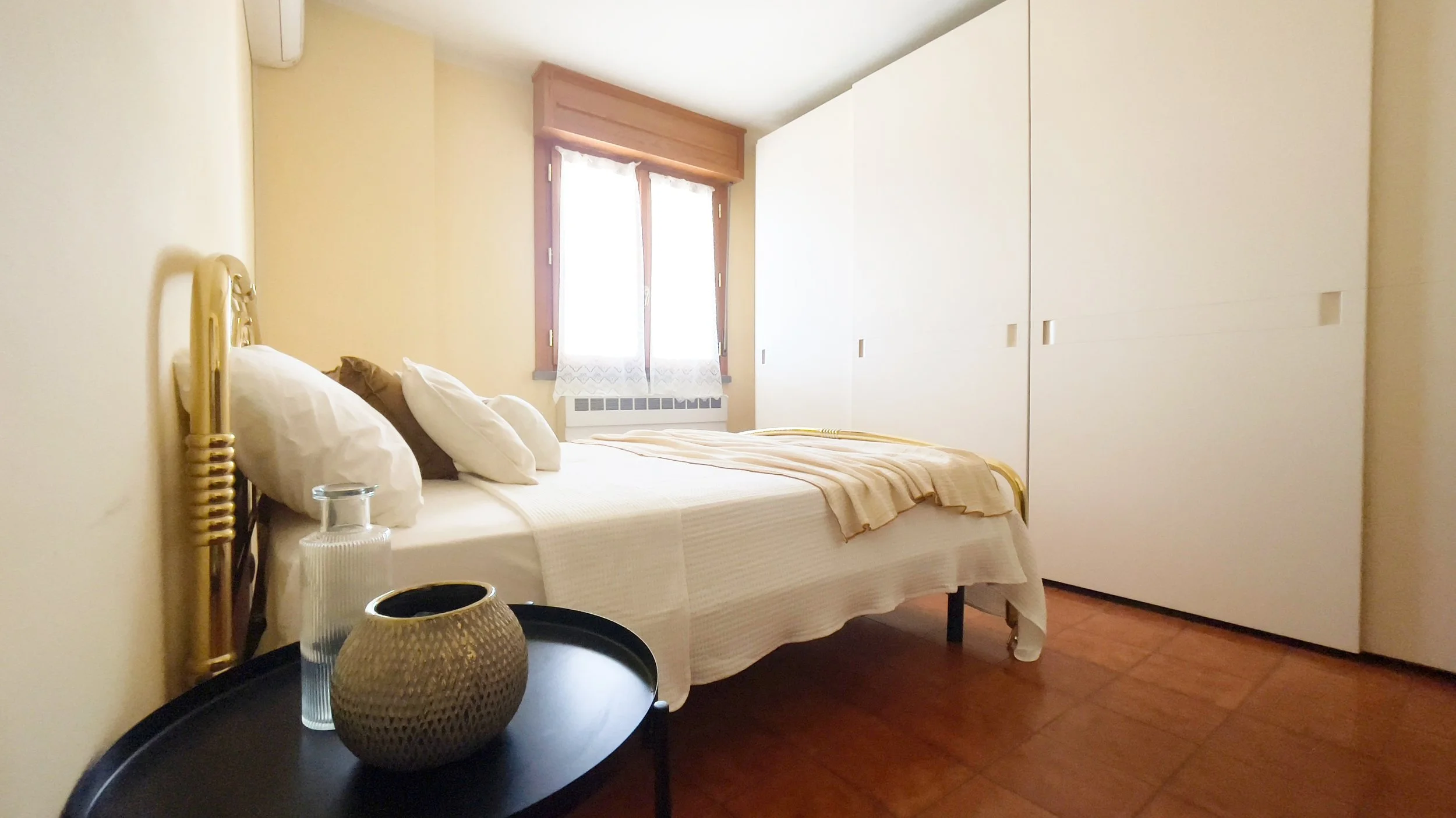 Camera da letto con letto matrimoniale, biancheria bianca, comodino nero, armadio bianco e finestra con tende in pizzo.
