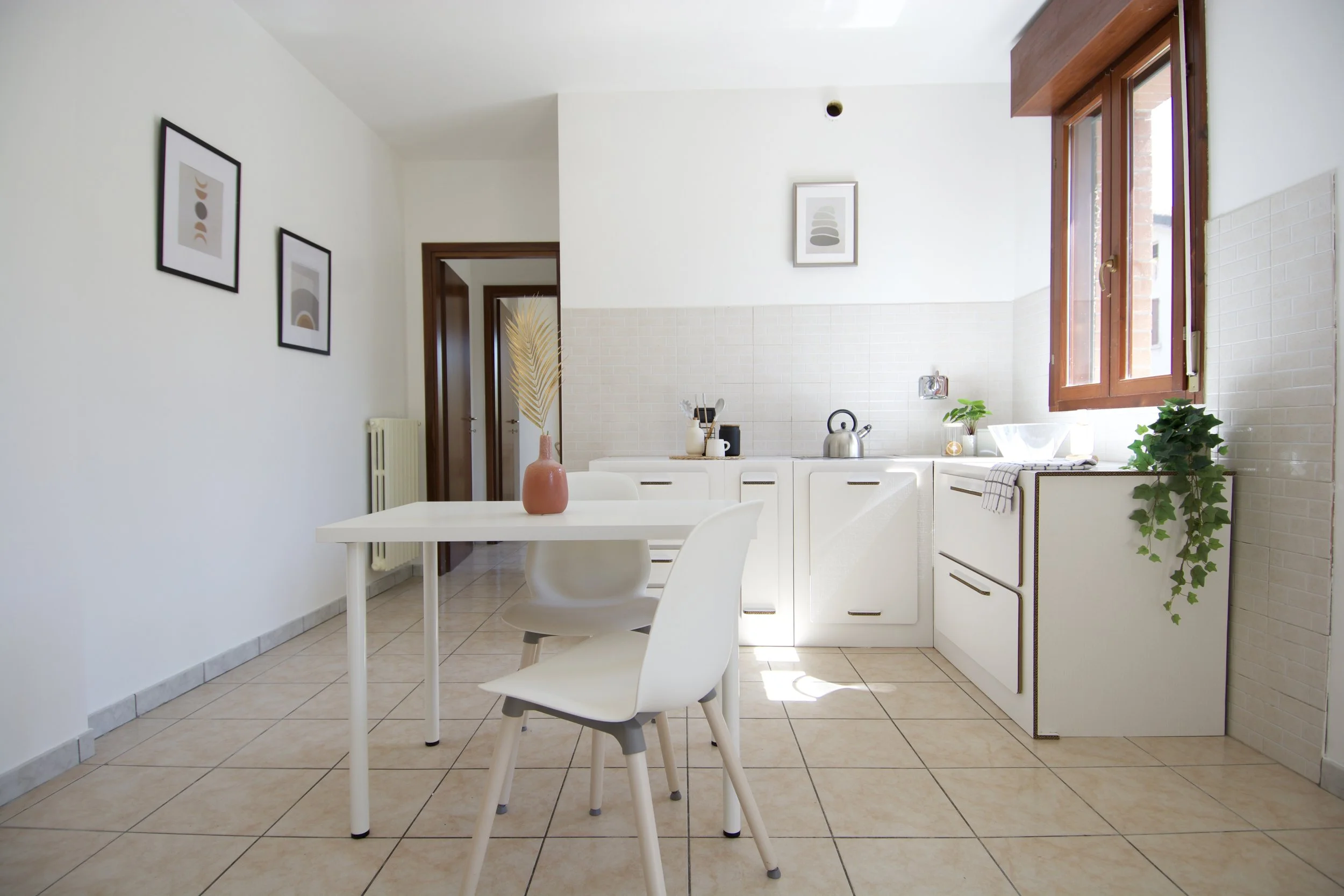 Cucina moderna e minimalista con tavolo bianco, sedia, decorazioni vegetali, mobilio chiaro, finestra con luce naturale, e quadri su parete.