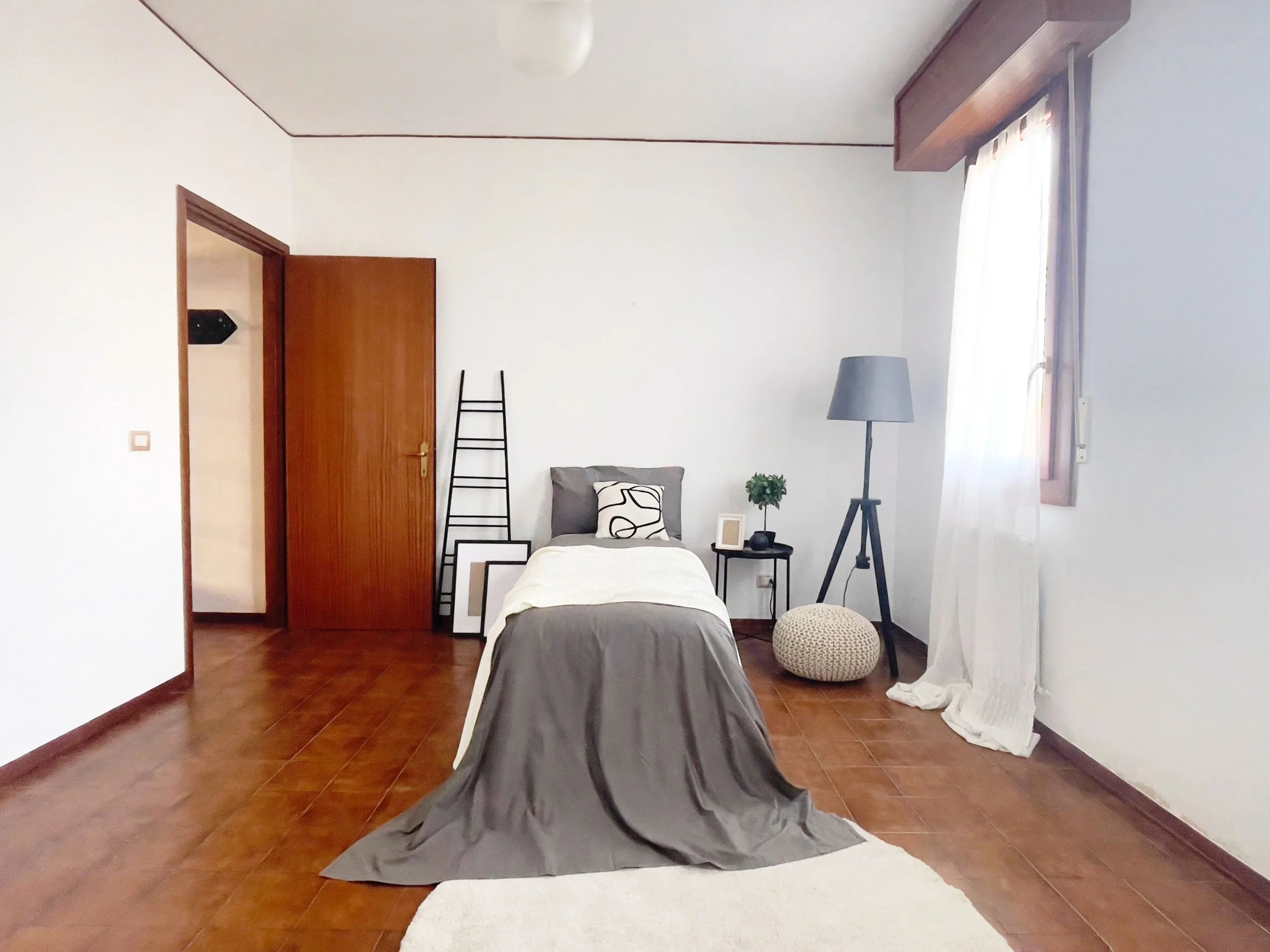 Camera da letto minimalista con letto singolo, parete bianca, pavimento in legno scuro, lampada da terra, cuscino grigio e finestra con tende bianche trasparenti.