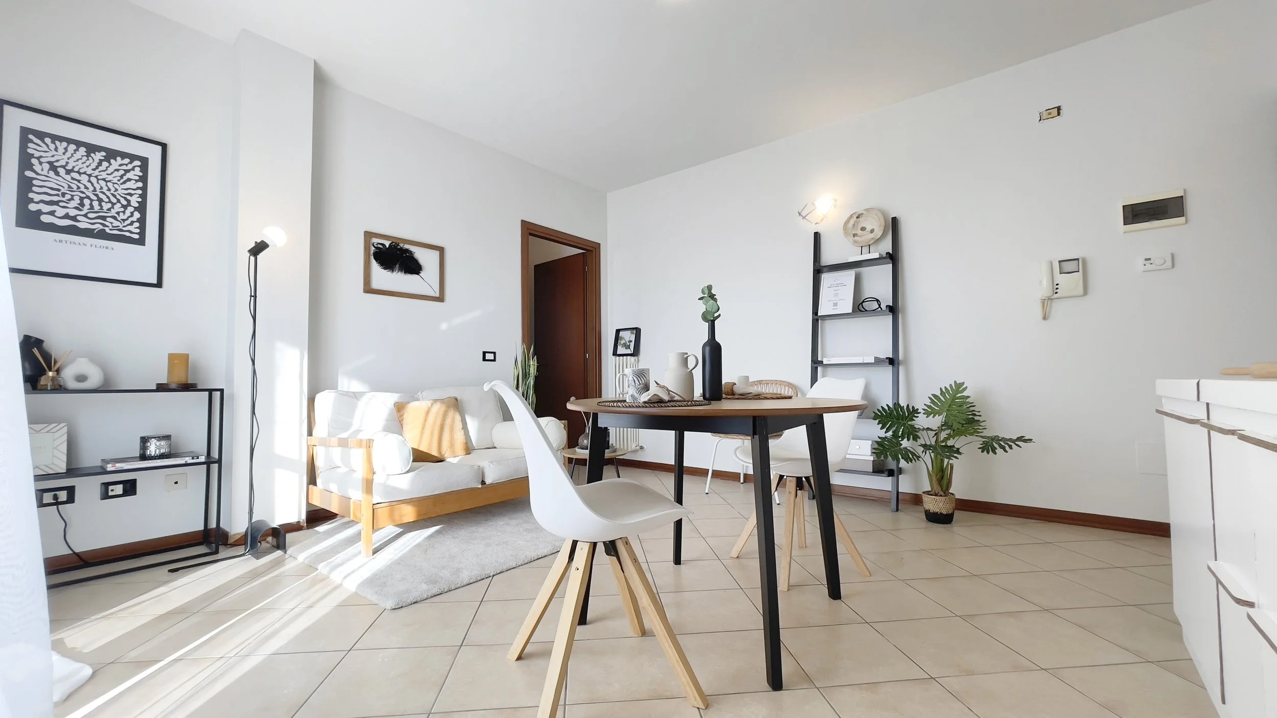 Soggiorno moderno e luminoso con arredamento minimalista; tavolo con sedie bianche, divano bianco, piante decorative, scaffali e quadri alle pareti.