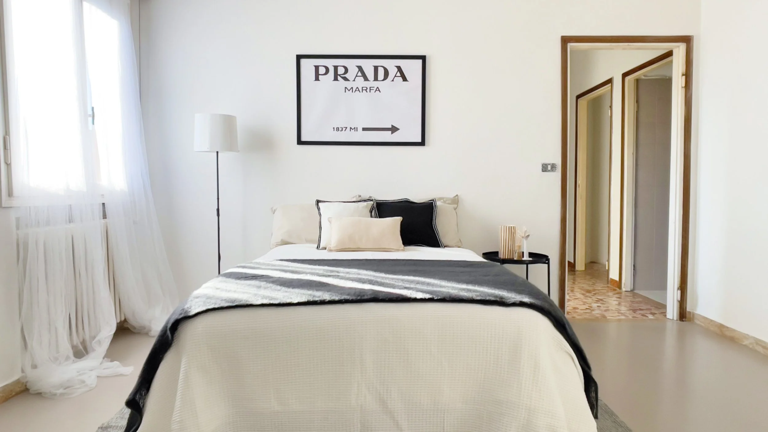 Camera da letto moderna con letto matrimoniale, cuscini chiari e scuri, lampada da terra, finestra con tende bianche, e quadro sul muro."