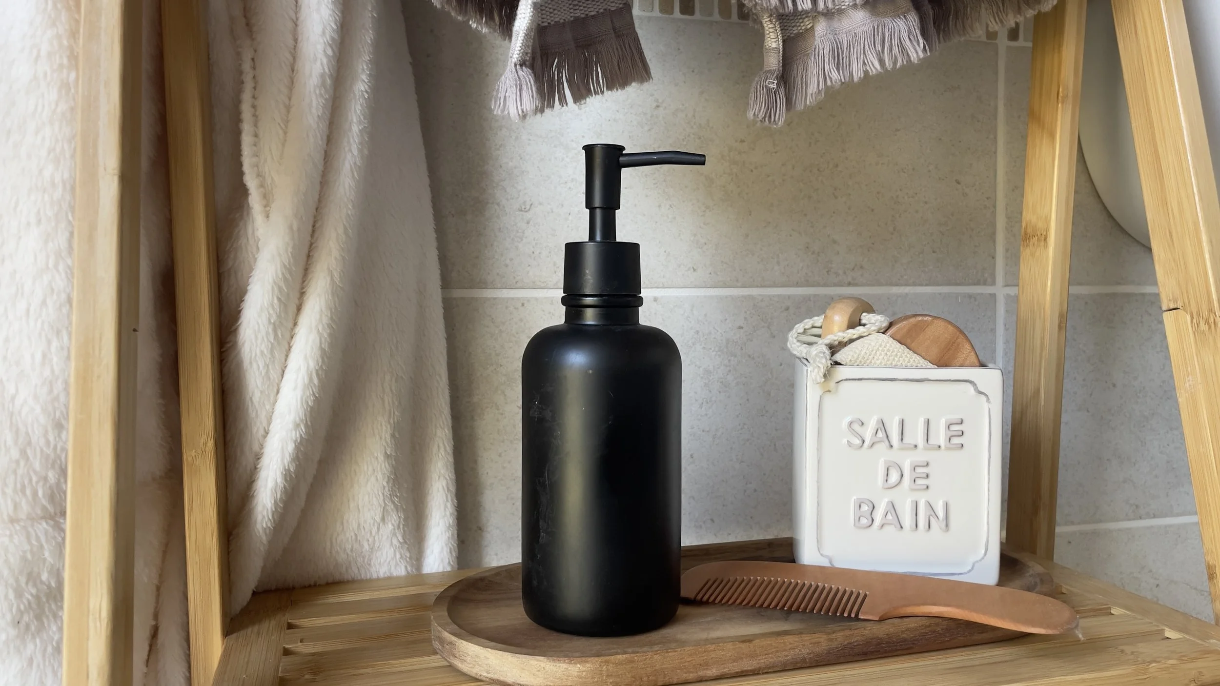 Dispensatore di sapone nero su un vassoio di legno con pettine, accanto a un contenitore con la scritta "SALLE DE BAIN" e accessori da bagno.