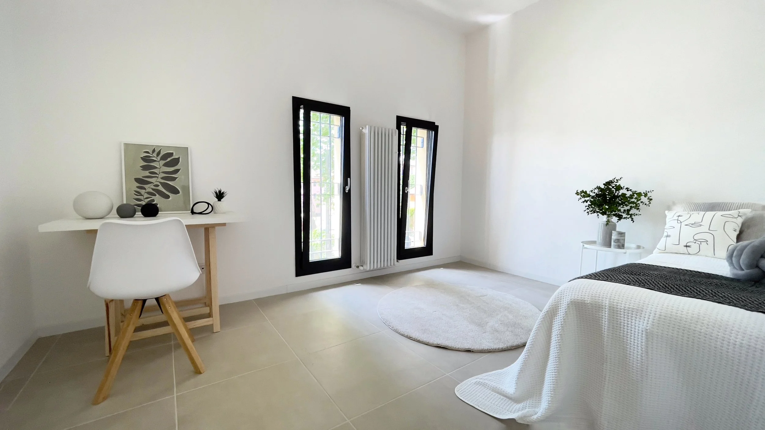 Camera moderna con pareti bianche, due finestre verticali, un piccolo tavolo con sedia bianca, decorazioni minimali e un letto con biancheria bianca e cuscino stampato.