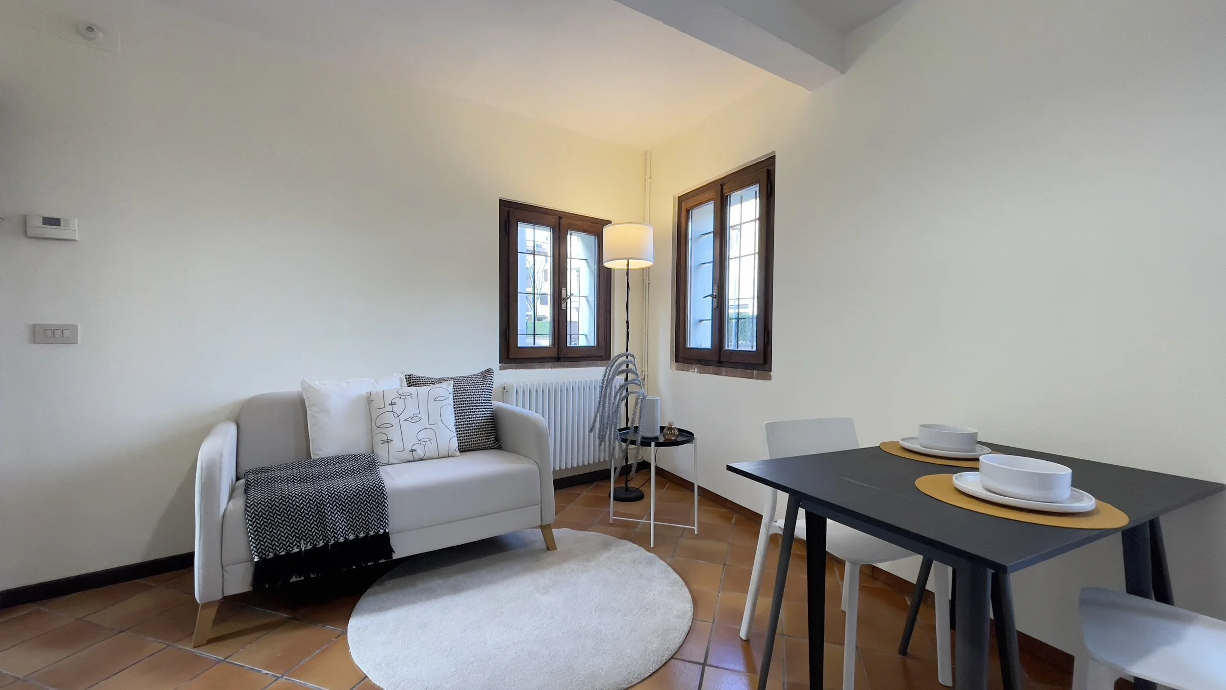 Salotto accogliente con divano bianco e cuscini decorativi, lampada a stelo, tappeto rotondo chiaro, tavolo con due posti apparecchiati per cena separati, pavimento in piastrelle marroni e finestra luminosa.