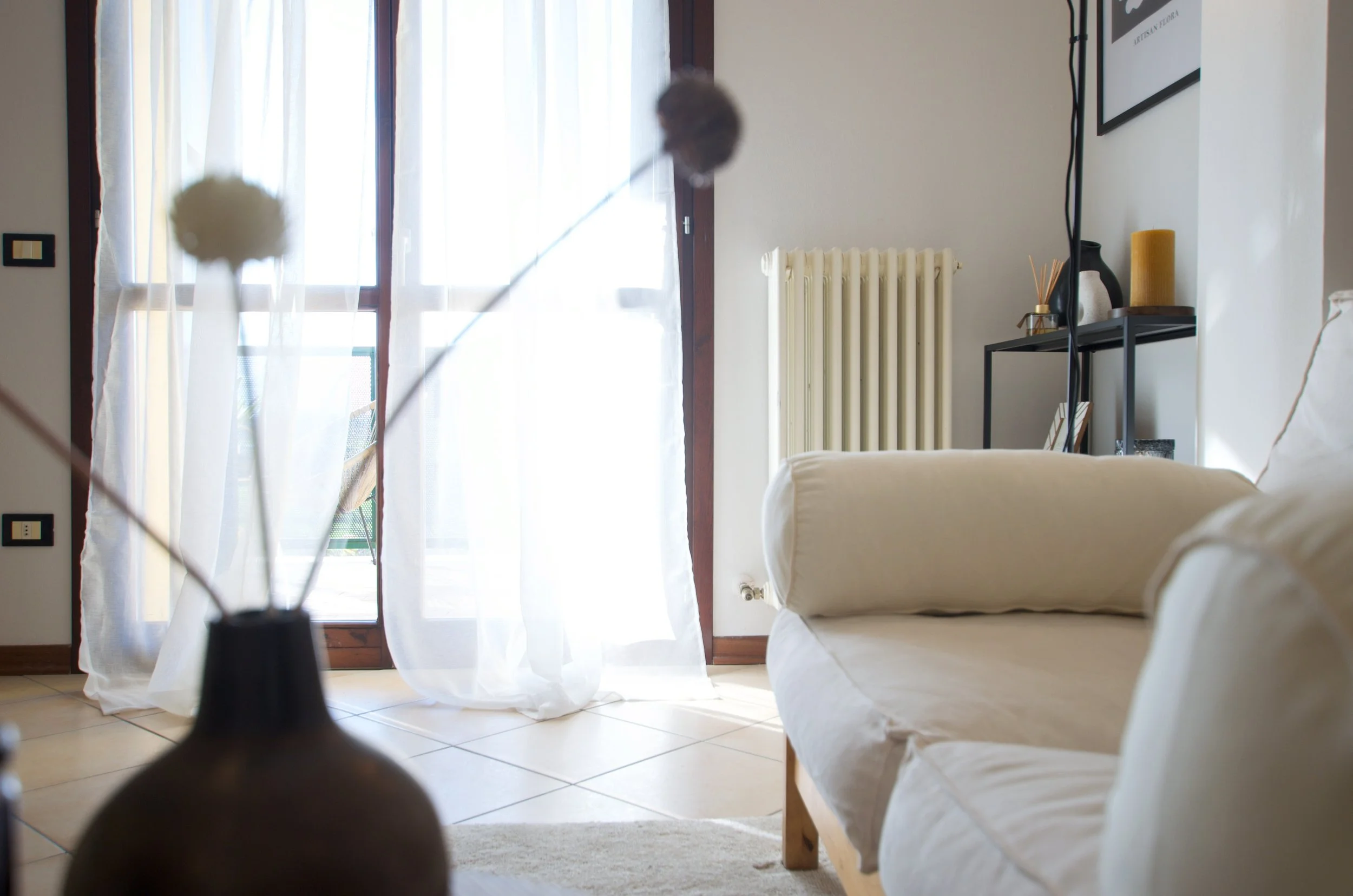 Soggiorno luminoso con divano bianco, finestra con tende bianche, radiatore e mensole con decorazioni.