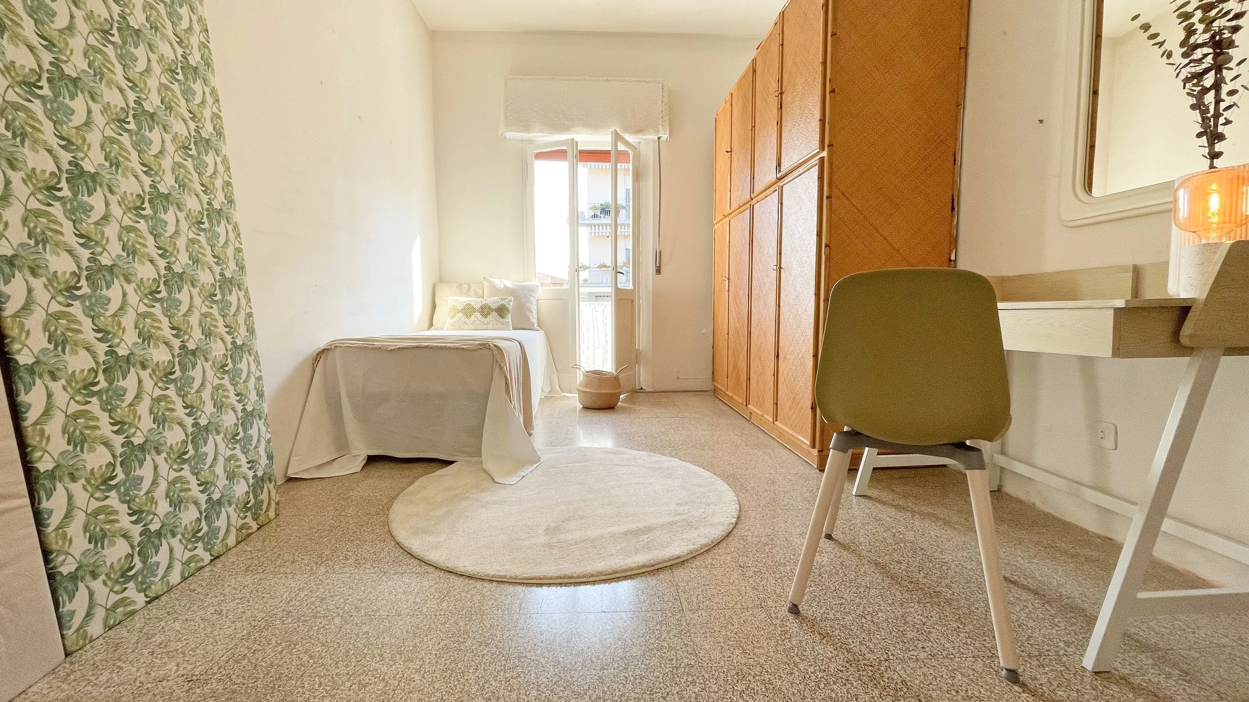 Camera da letto luminosa con letto singolo, tappeto rotondo chiaro, armadio in legno, scrivania con sedia, e finestra aperta che lascia entrare la luce del sole.