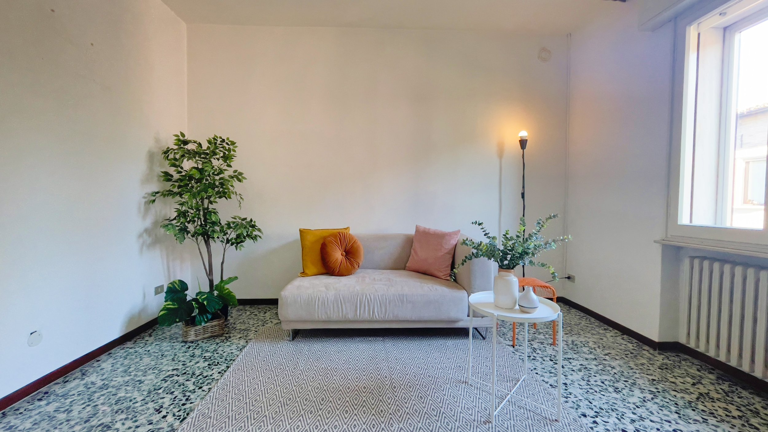 Soggiorno moderno con divano beige, cuscini colorati, tavolino bianco con vasi, pianta decorativa, lampada e finestra luminosa.
