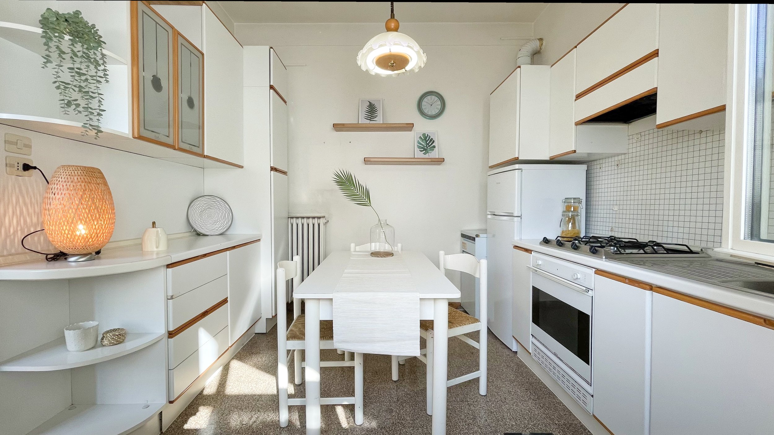 Cucina moderna con mobili bianchi, tavolo con sedie bianche, pianta decorativa, lampada a sospensione, fornello e forno, frigorifero e decorazioni minimaliste sulle mensole.