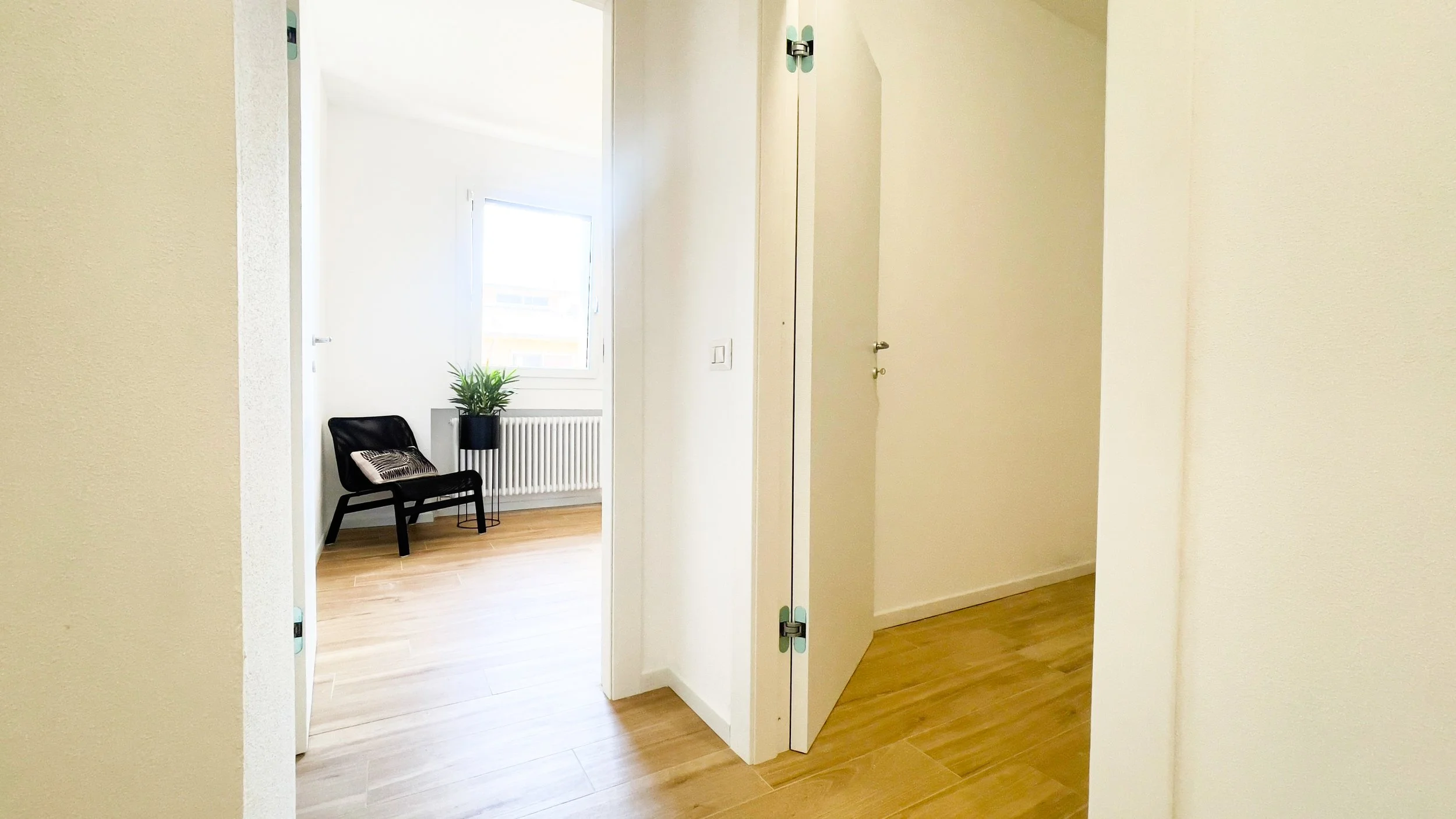 Corridoio di un appartamento moderno con pavimento in legno, una porta aperta, una sedia nera e una pianta in vaso.