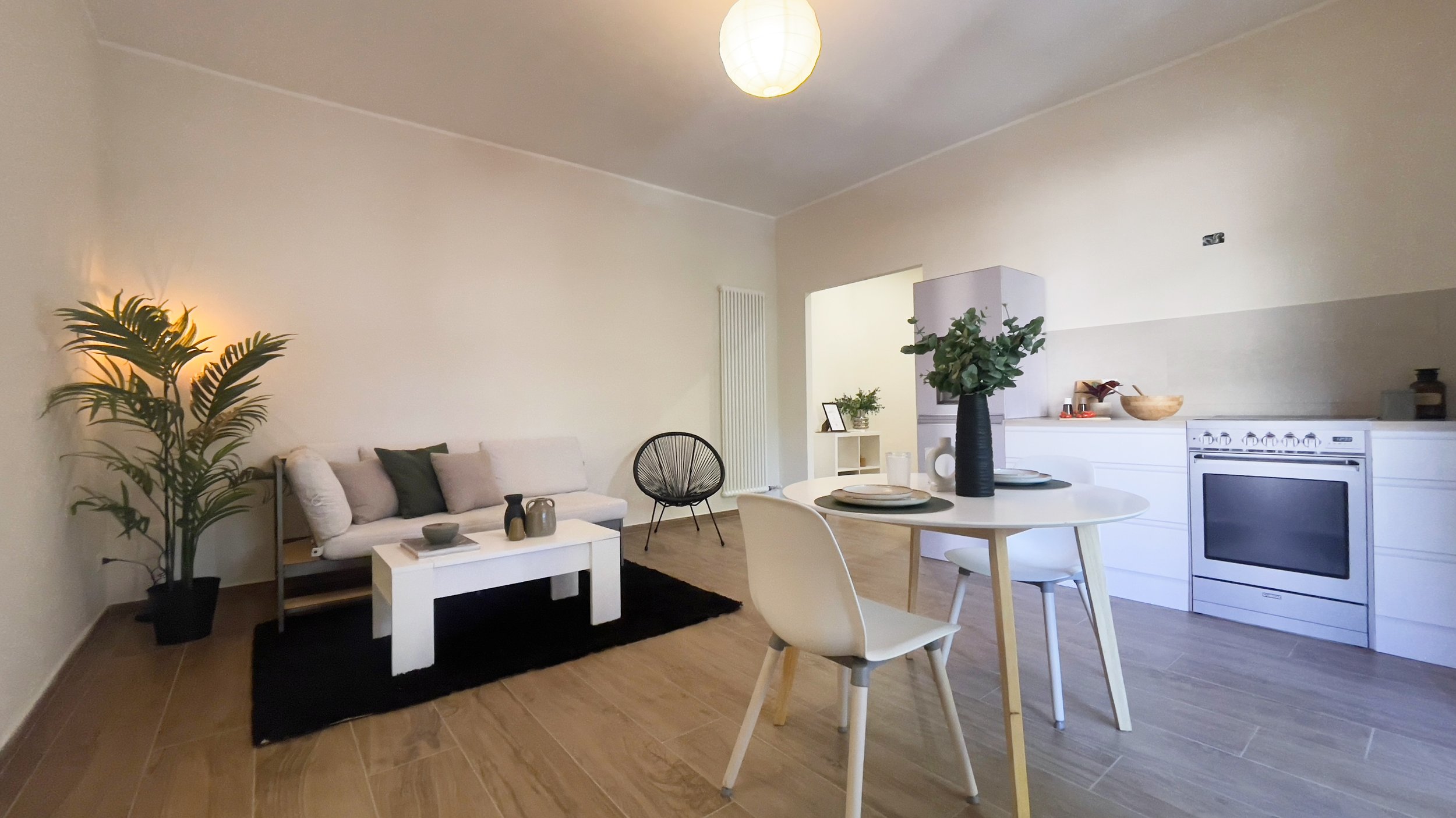 Soggiorno moderno con divano, tavolino, sedia e piante, cucina integrata con forno e tavolo da pranzo.