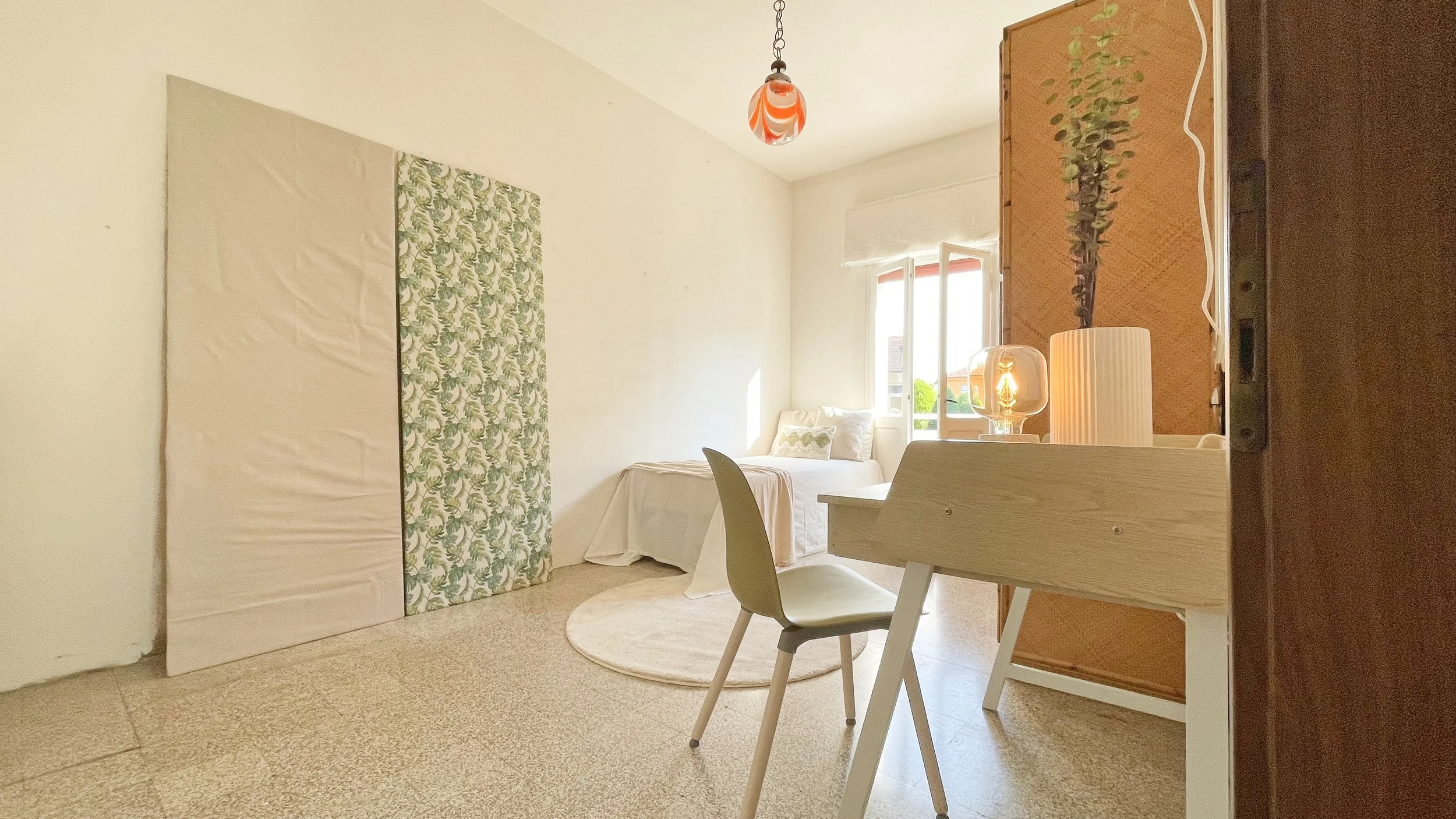 Camera da letto minimalista con un letto singolo, sedia bianca e scrivania in legno chiaro. Decorazioni murali astratte, lampada ornamentale e finestra aperta con luce naturale. Atmosfera luminosa e accogliente.