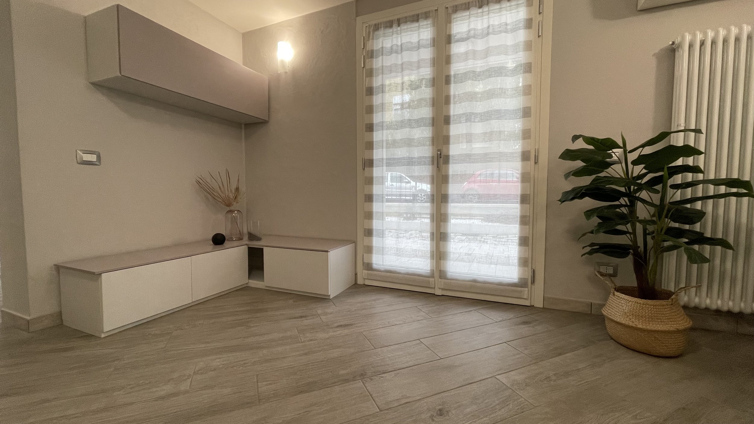 Interno di un soggiorno moderno con pavimento in legno chiaro, mobile basso bianco, pianta in vaso di vimini, e porta finestra con tendaggi a righe.