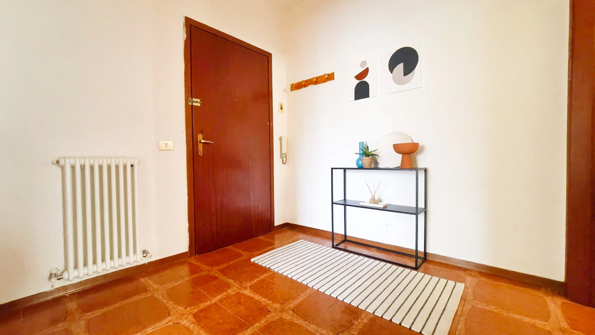 Ingresso con porta marrone, radiatore bianco, piccolo tavolino nero con soprammobili, e quadro astratto sul muro.