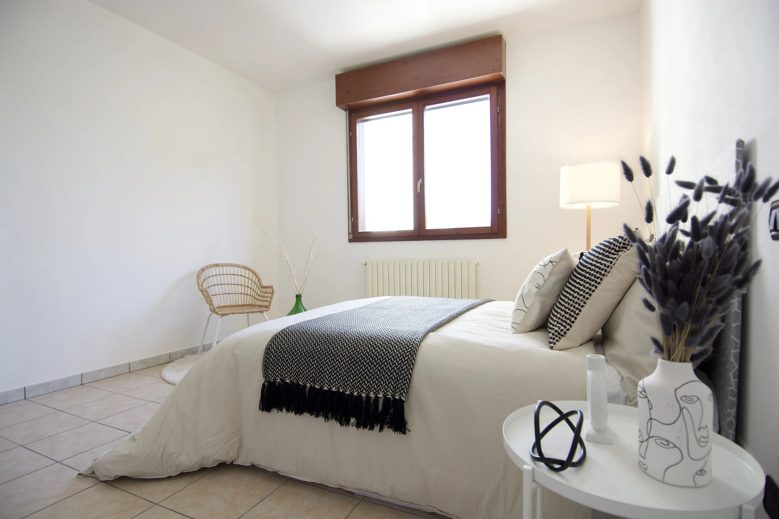 Camera da letto minimalista con letto matrimoniale, finestra con cornice in legno, sedia in vimini, comodino rotondo e decorazioni sobrie. Cuscini e copriletto in bianco e nero, lampada da tavolo e vaso con fiori secchi.