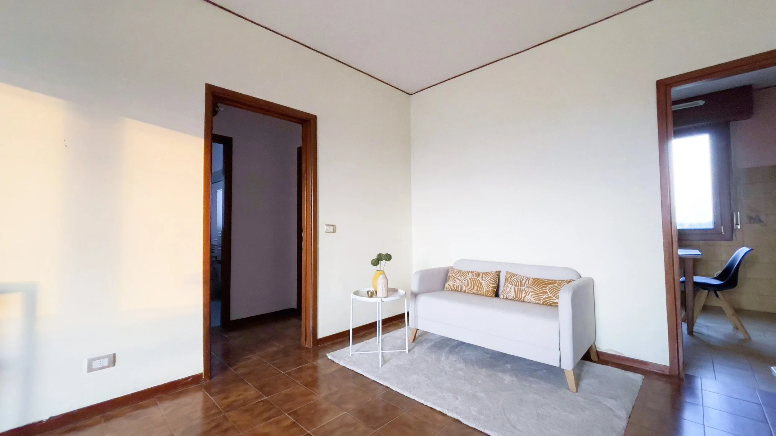 Soggiorno minimalista con divano bianco, cuscini decorativi gialli, tavolino bianco con pianta, tappeto grigio, pavimento in piastrelle marroni e due porte per altre stanze.