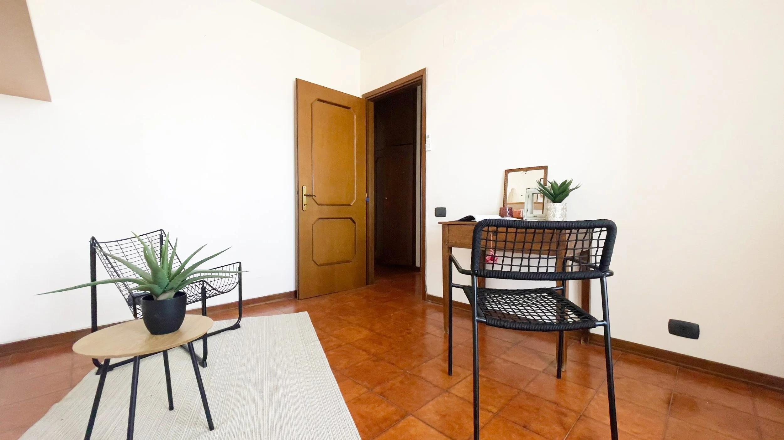 Interno di una stanza con una sedia in metallo nero, un tavolino con pianta in vaso, una scrivania con specchio e piante, e una porta aperta su un pavimento in piastrelle marroni.