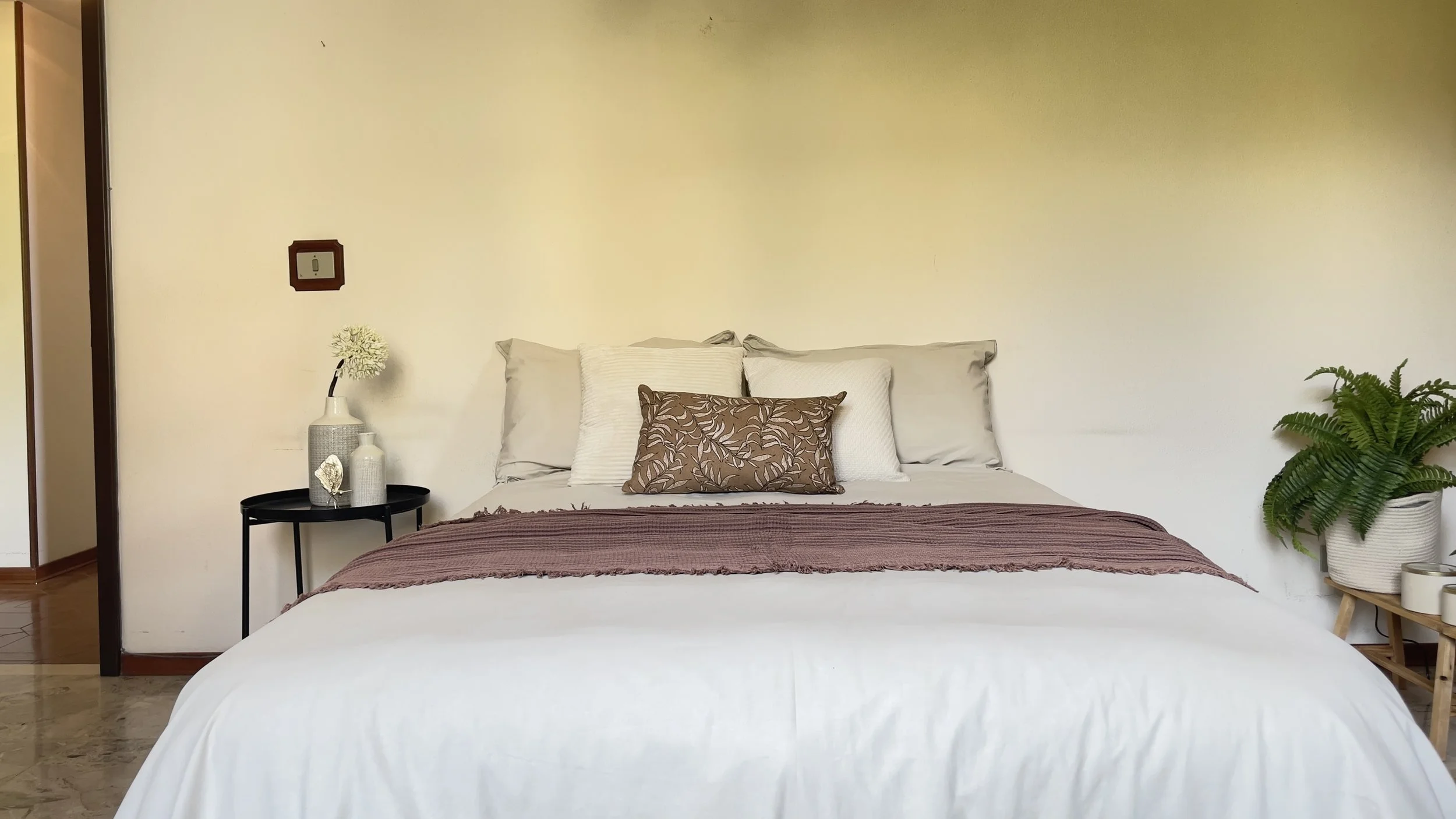 Camera da letto minimalista con letto matrimoniale, cuscini decorativi, un tavolino con vasi e pianta verde a destra.