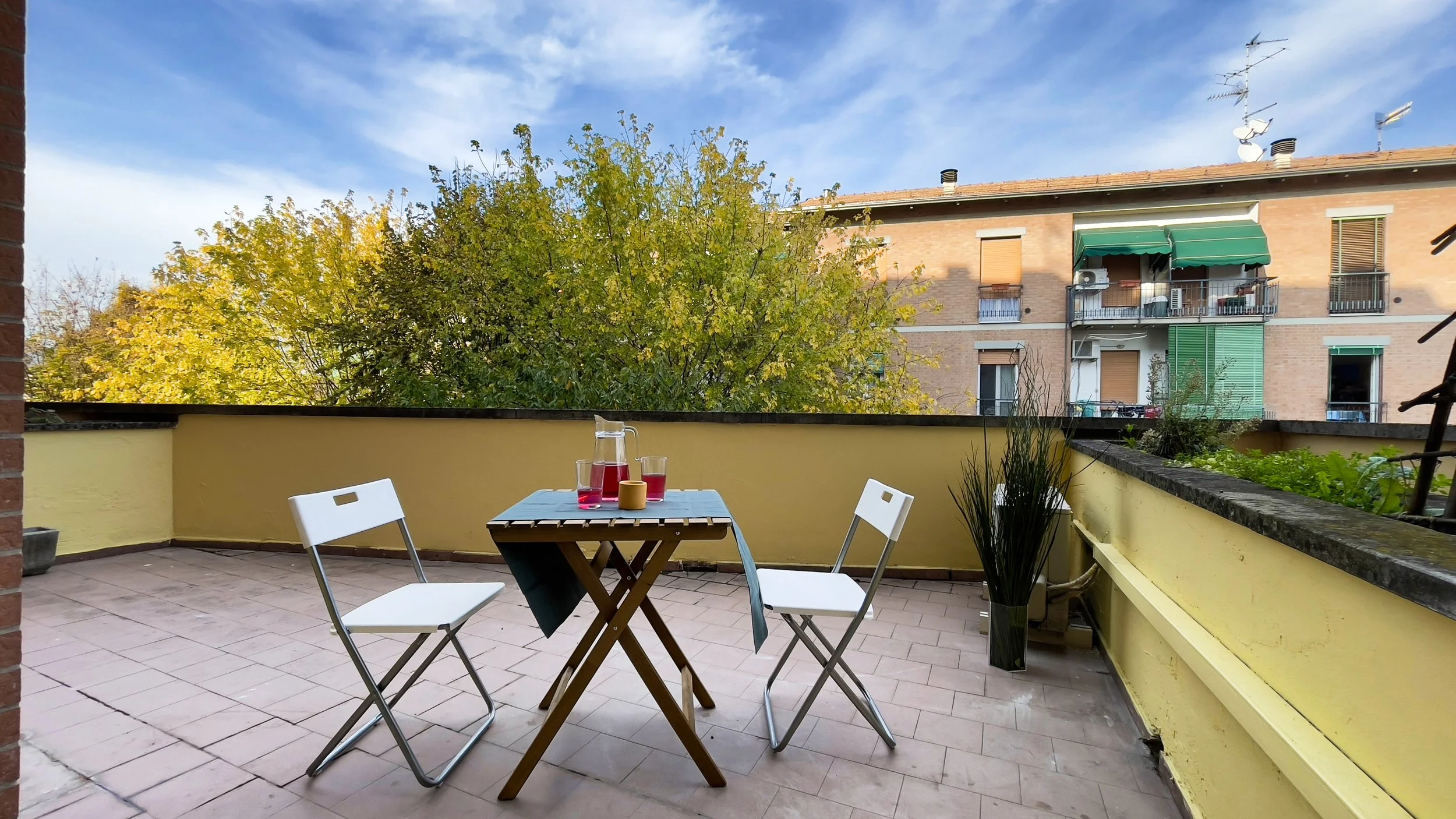 Terrazza con tavolo, sedie, caraffa con bevanda rossa e bicchieri; edificio e alberi sullo sfondo.