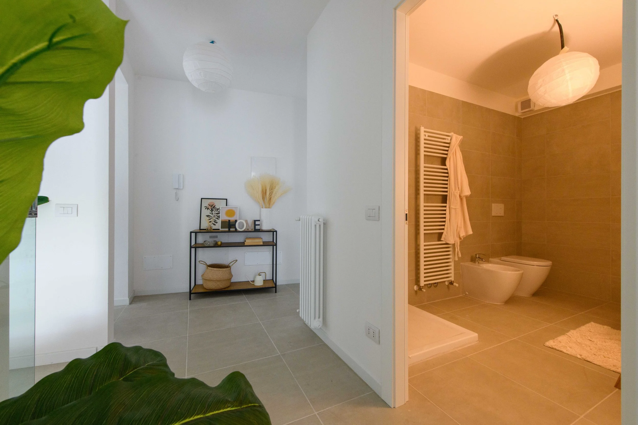 Ingresso di una casa con un bagno sulla destra. La stanza è arredata con tonalità neutre, piante verdi e un mobiletto decorato. Una porta aperta mostra un bagno con sanitari moderni e un portasciugamani riscaldato.