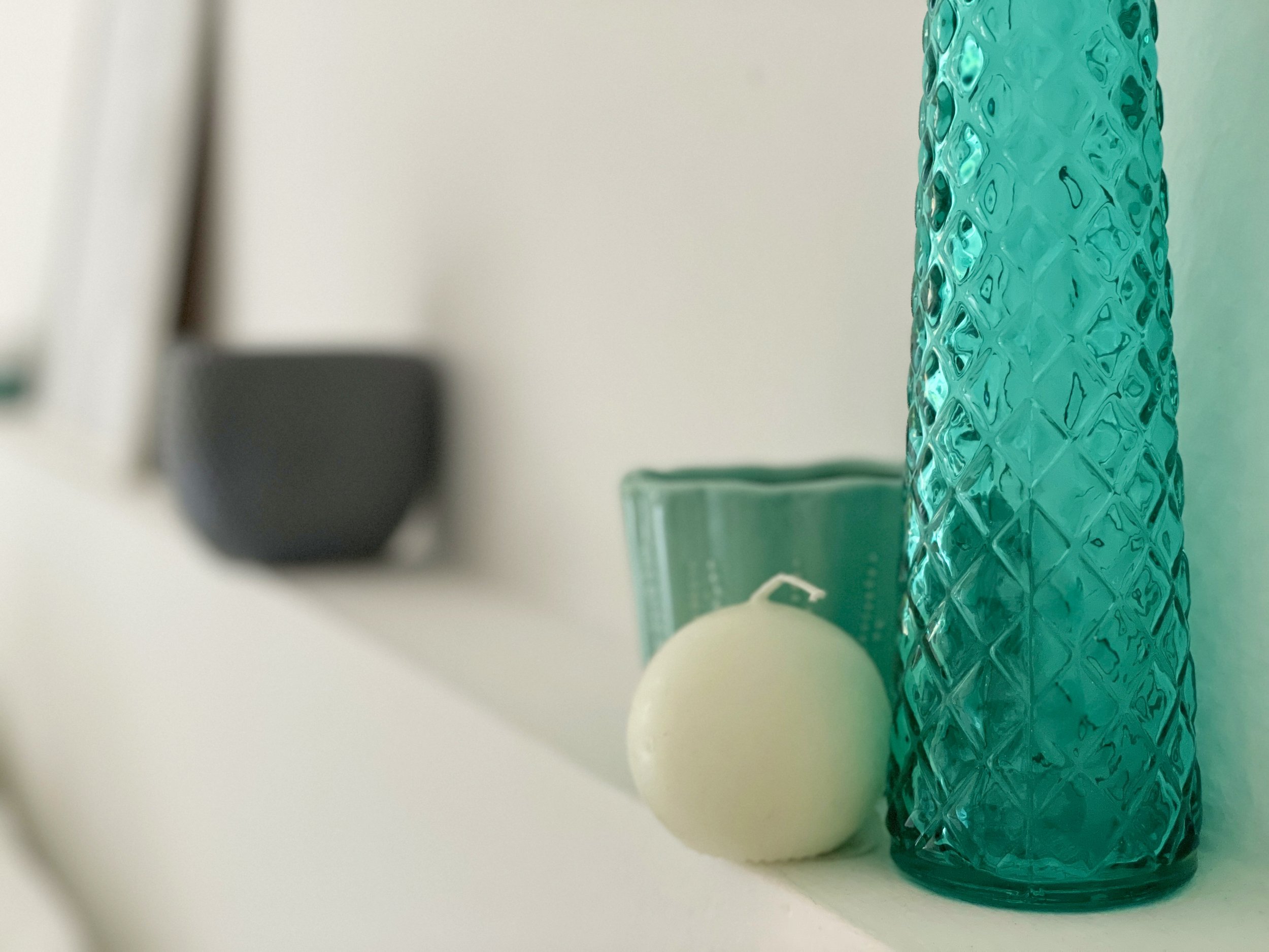 Bottiglia decorativa in vetro trasparente verde, candela bianca rotonda e tazza in ceramica verde su una mensola bianca.