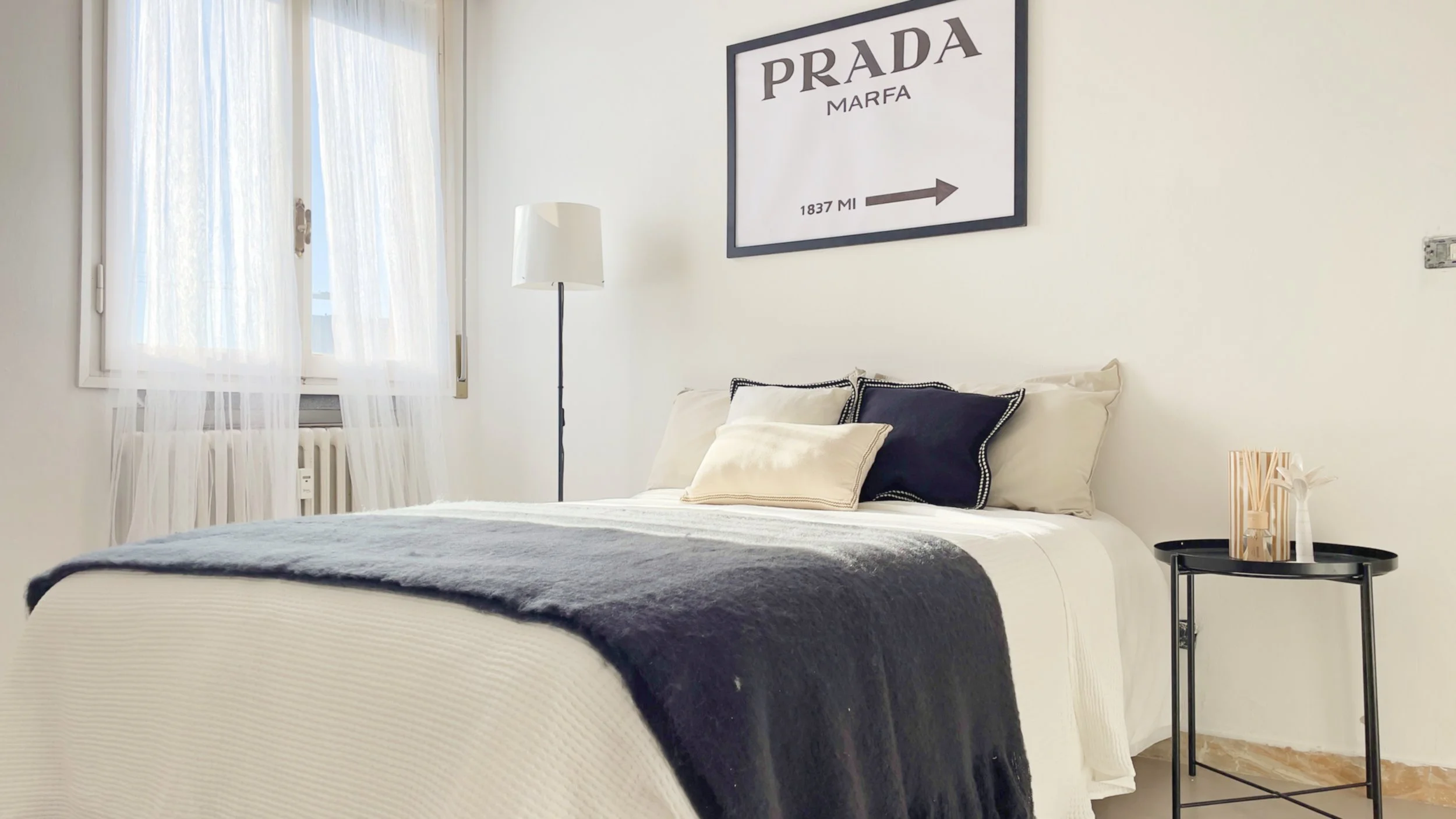 Camera da letto moderna con letto matrimoniale, cuscini decorativi, coperta scura e tavolino nero; una finestra con tende bianche e un quadro "PRADA MARFA" sopra il letto.