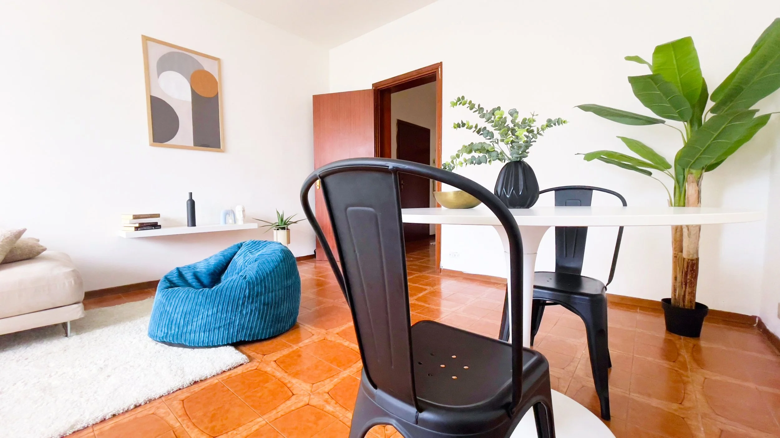 Soggiorno moderno con sedia nera, tavolo bianco, pianta verde, pouf blu, tappeto bianco e decorazione a parete astratta.