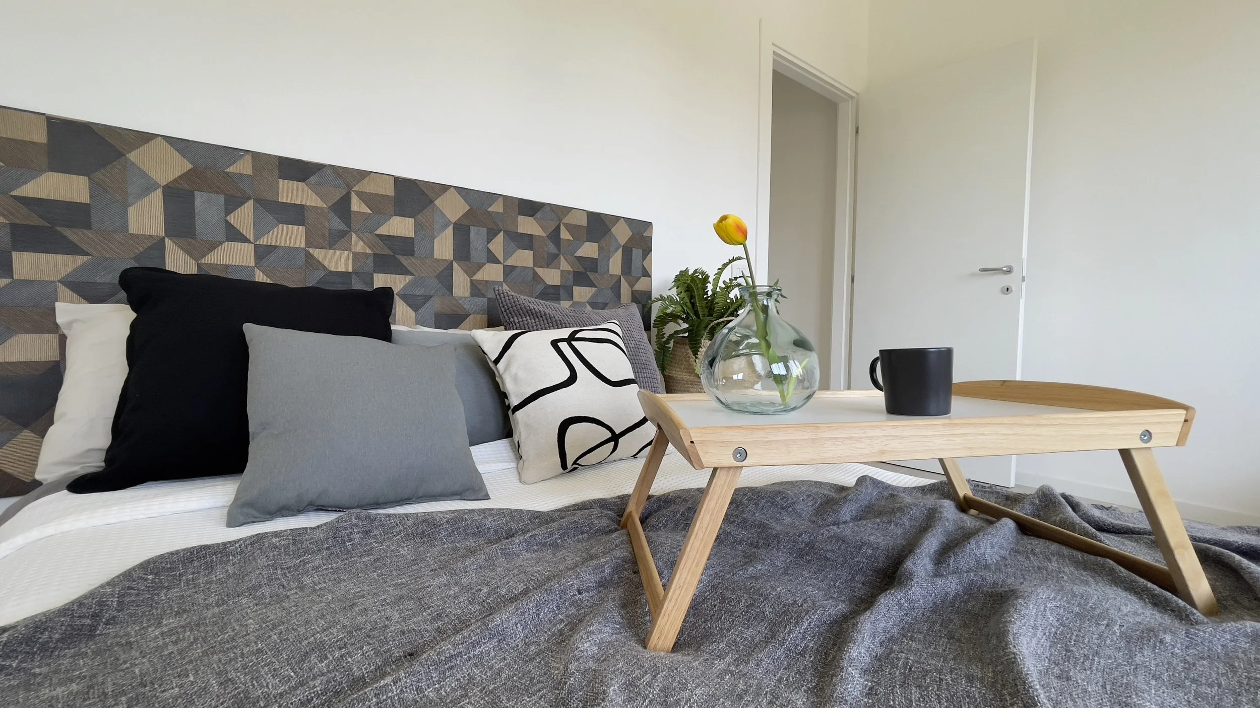 Interno di una camera da letto moderna con letto matrimoniale, testata geometrica, cuscini variopinti, vassoio di legno con tazza e vaso con fiore giallo.