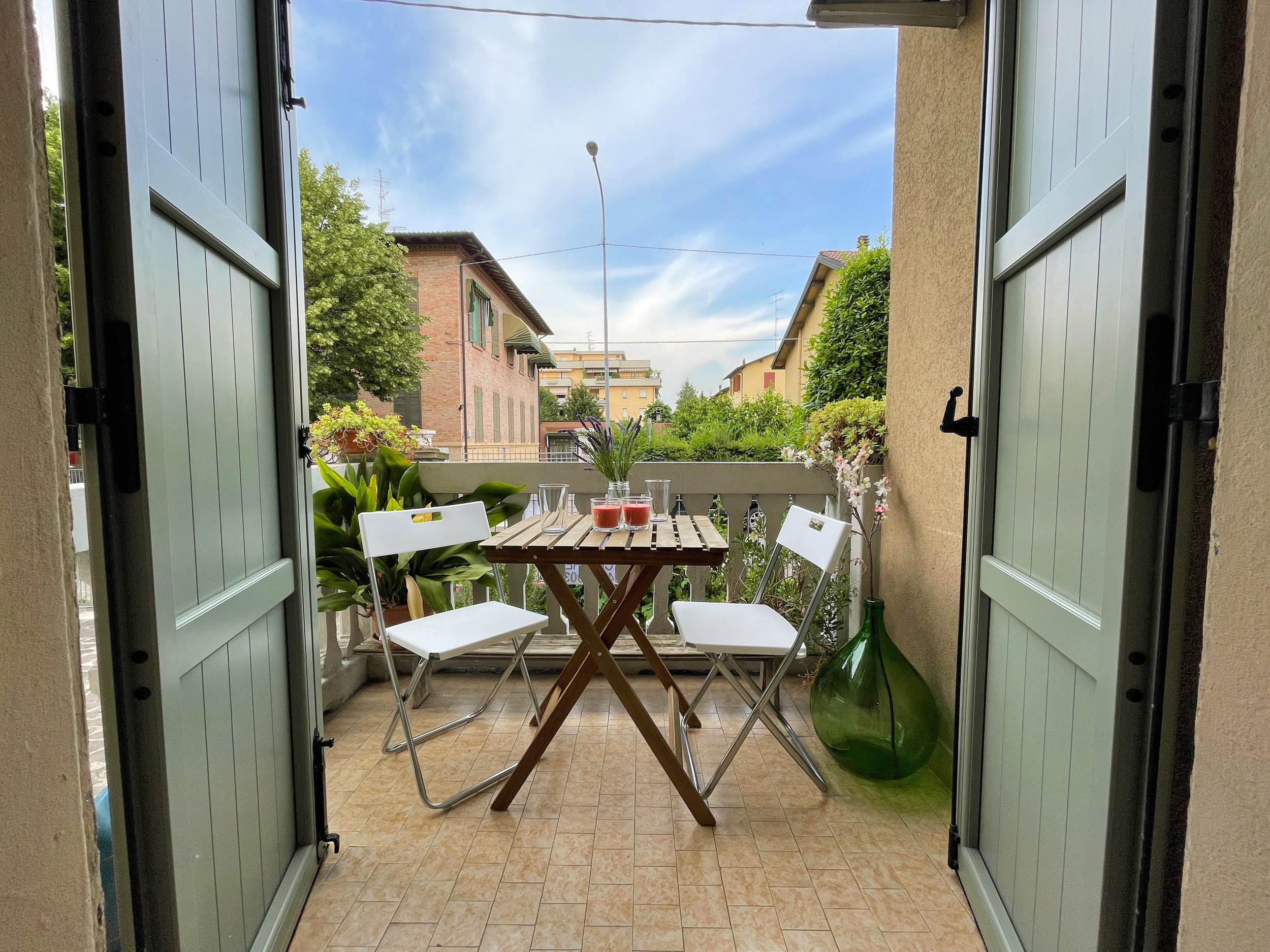 Piccolo balcone con tavolo e sedie pieghevoli bianche, bevande sul tavolo, piante decorative, e fiori sul parapetto, vista su edifici residenziali.