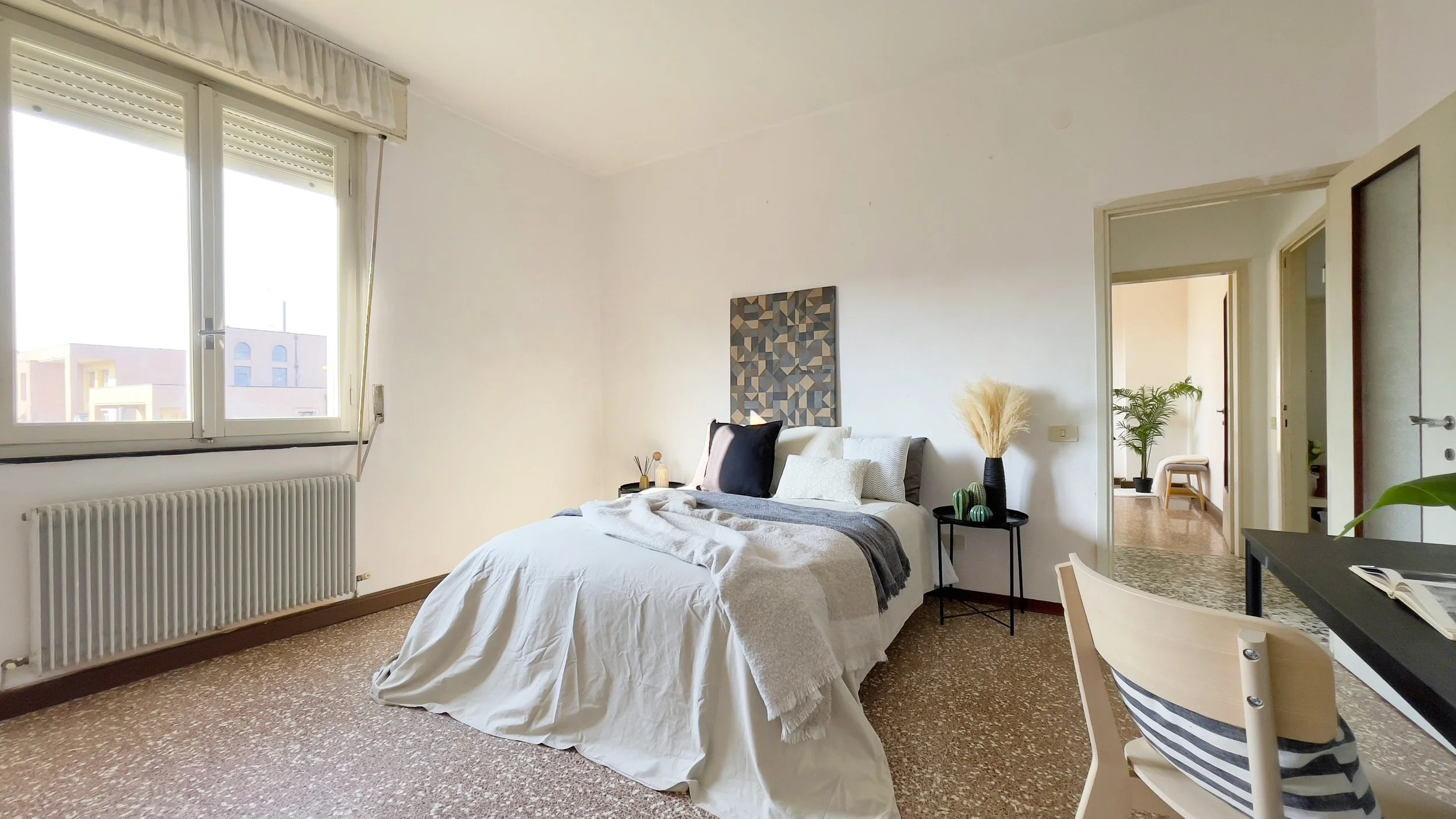 Camera da letto luminosa con letto matrimoniale, lenzuola bianche, cuscini decorativi e coperta grigia. Tavolino nero con vaso e piante. Finestra grande, pavimento in marmo. Arredo moderno e minimalista.