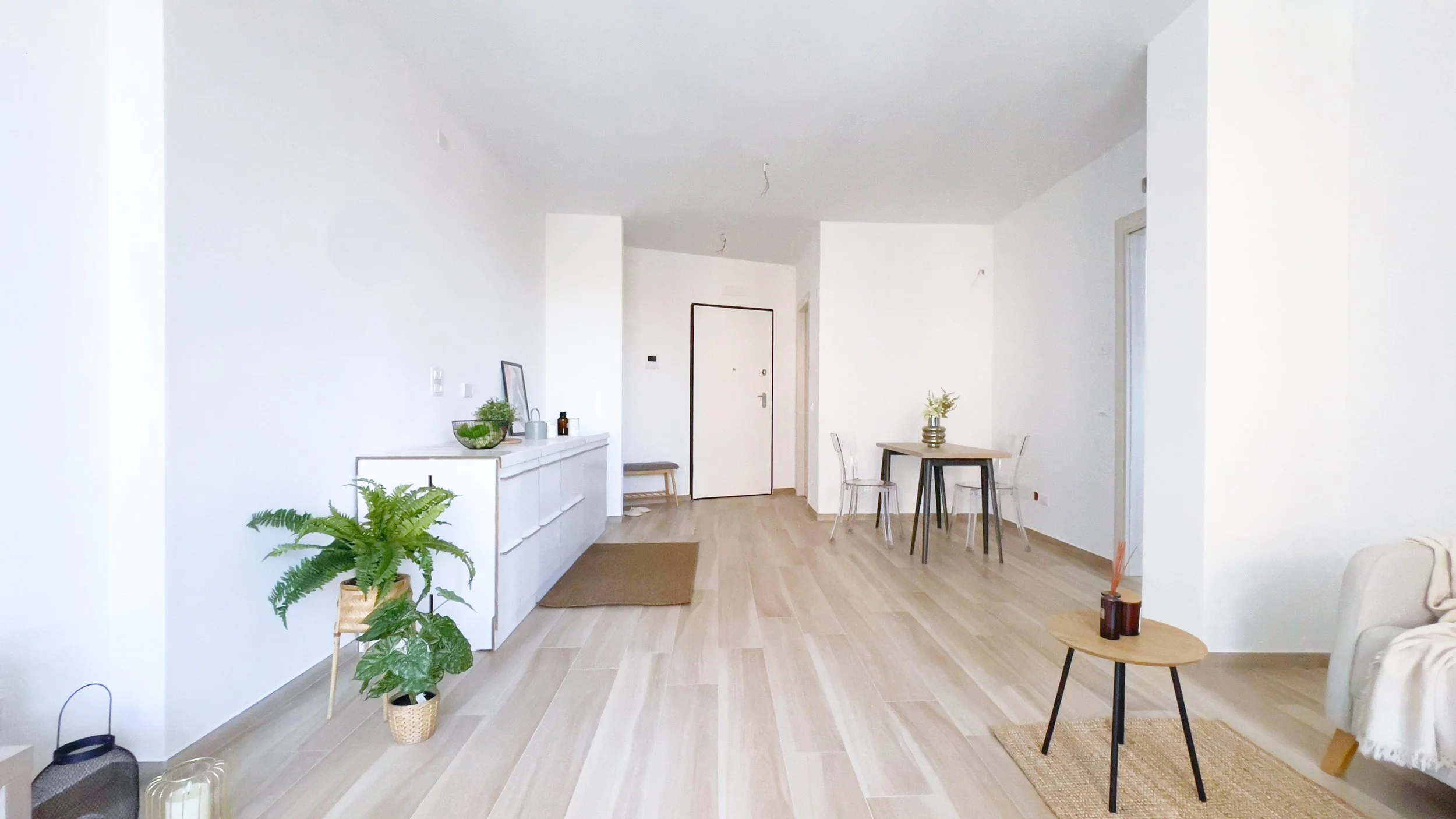 Interno di un luminoso soggiorno moderno con pavimento in legno chiaro. Ci sono piante in vaso, un mobile bianco, un tavolino con candele e una sedia trasparente accanto a un tavolo più grande. La porta d'ingresso è visibile sullo sfondo.