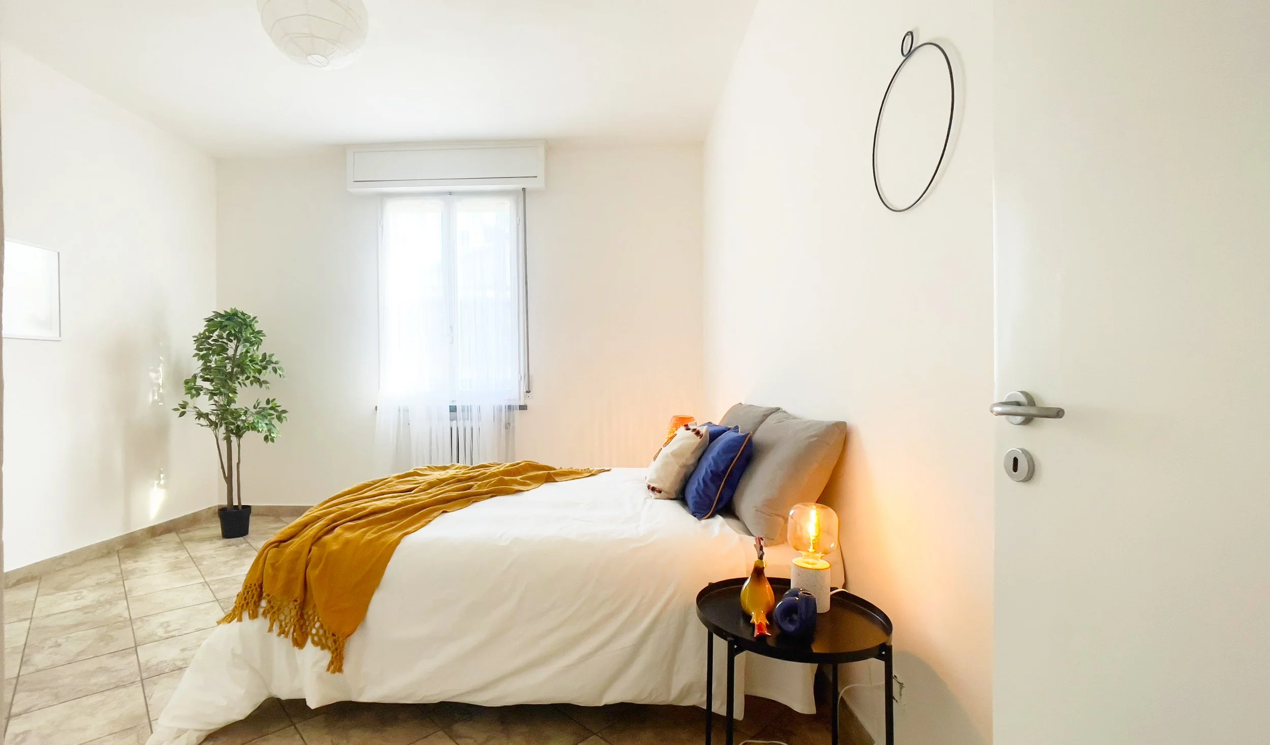 Camera da letto moderna e luminosa con letto matrimoniale, coperte bianche e gialle, cuscini colorati, un comodino con lampada e decorazioni, pianta in un angolo e finestra con tende bianche.