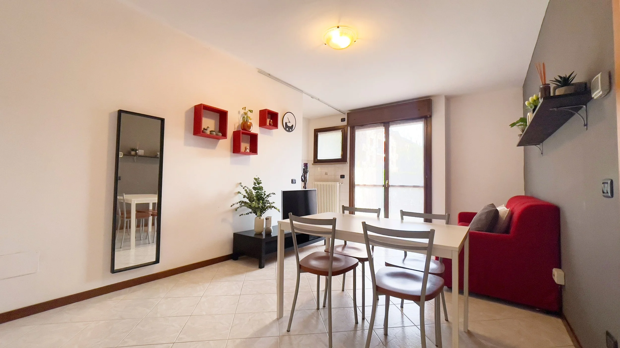 Soggiorno moderno con tavolo e sedie bianche, specchio inclinato, scaffale rosso a parete, divano rosso e pianta decorativa, finestra grande.
