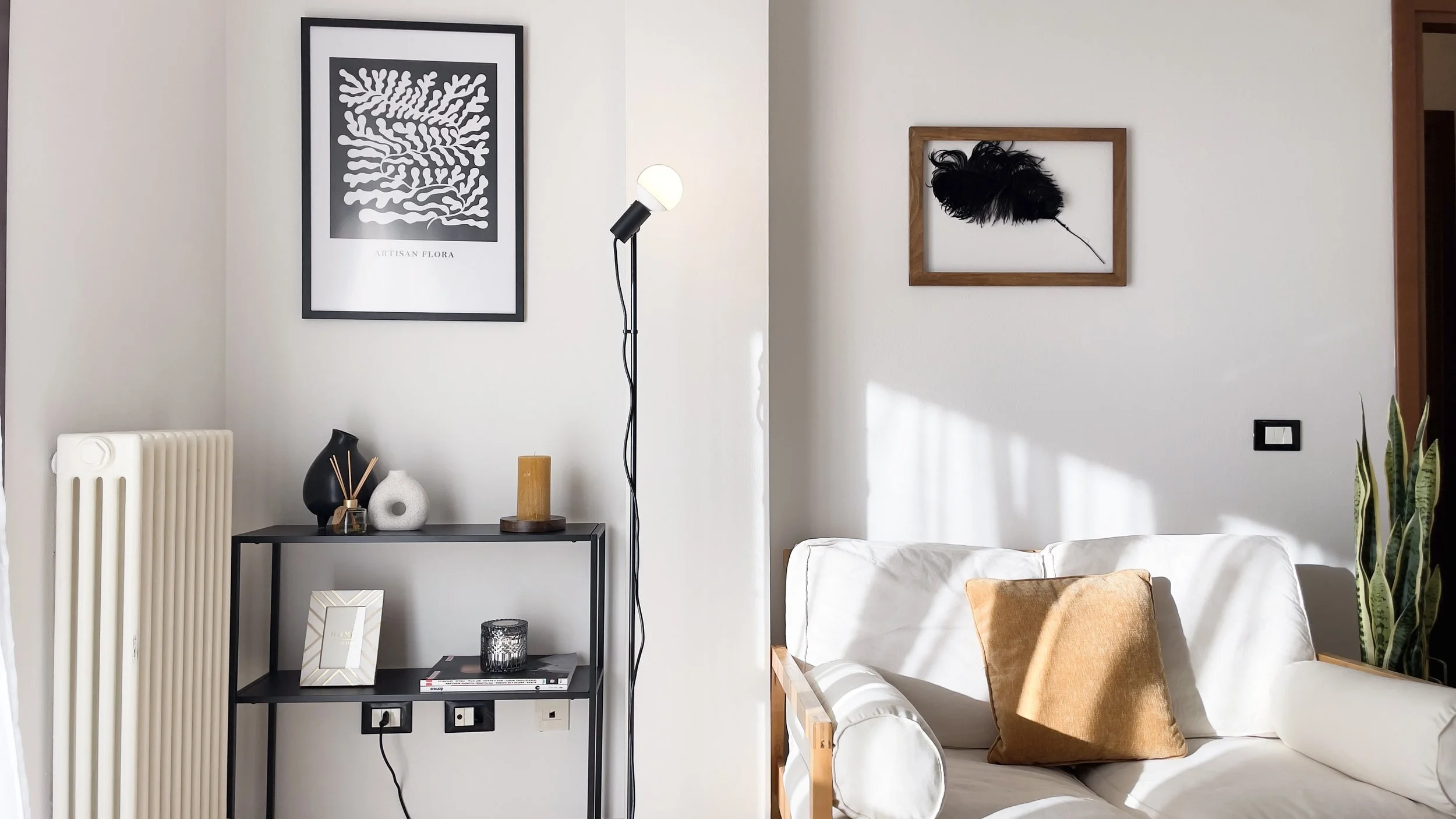 Soggiorno moderno con divano bianco, lampada da terra nera, mensola con vasi e oggetti decorativi, quadri alle pareti, e pianta in un angolo.