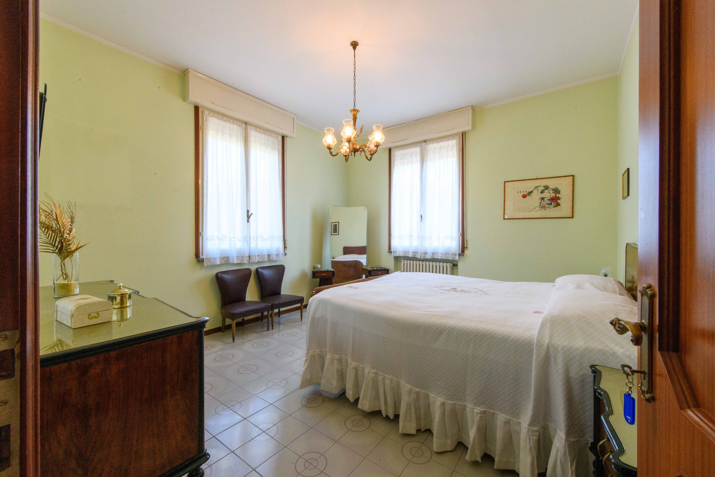Camera da letto con letto matrimoniale, lampadario, due finestre con tende bianche, specchio, due sedie e comò con oggetti decorativi.