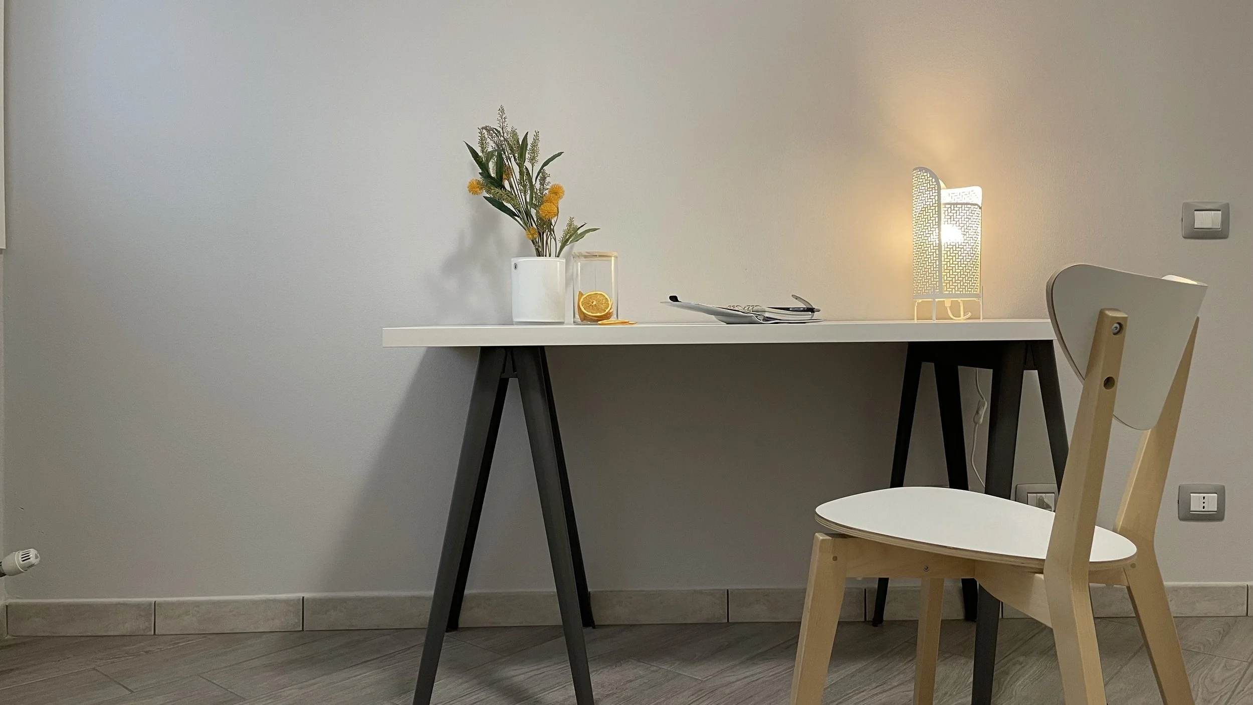 Tavolo con sedia in legno, vaso di fiori, bottiglia d'acqua con fetta d'arancia, riviste e lampada accesa.