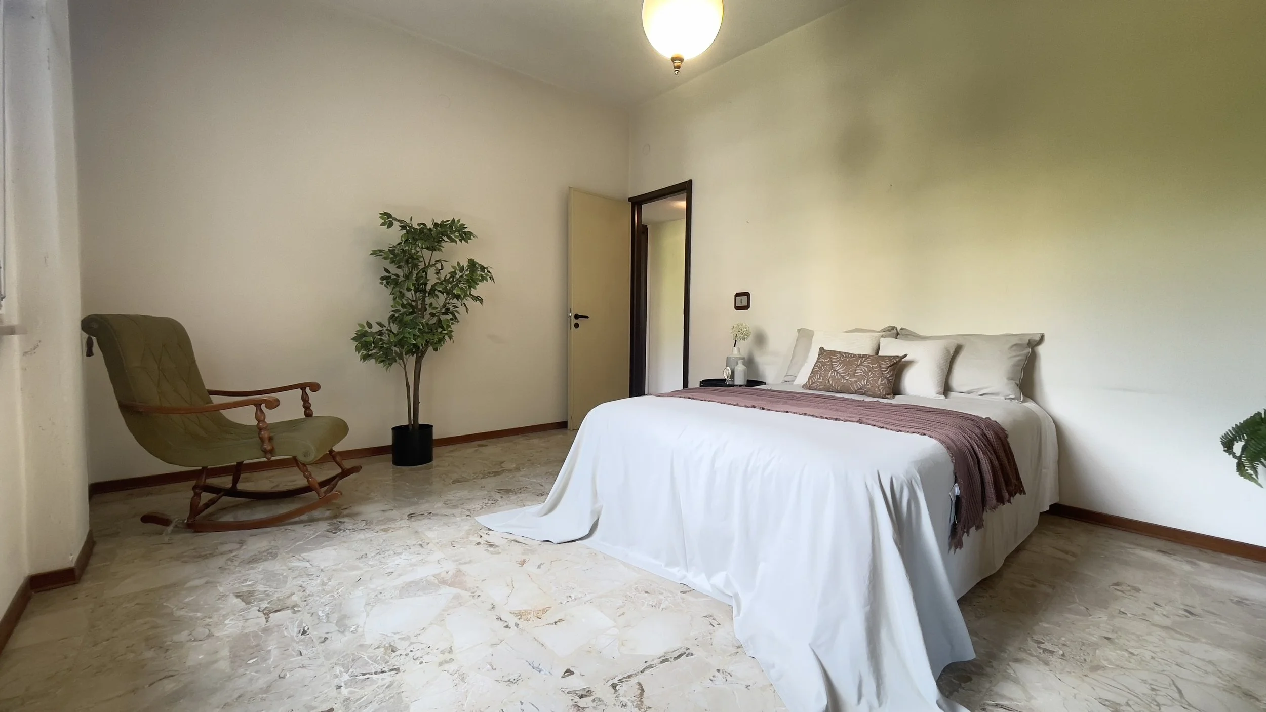 Camera da letto minimale con letto matrimoniale, copriletto bianco, cuscini decorativi, pianta in vaso, sedia a dondolo e lampada a sospensione.