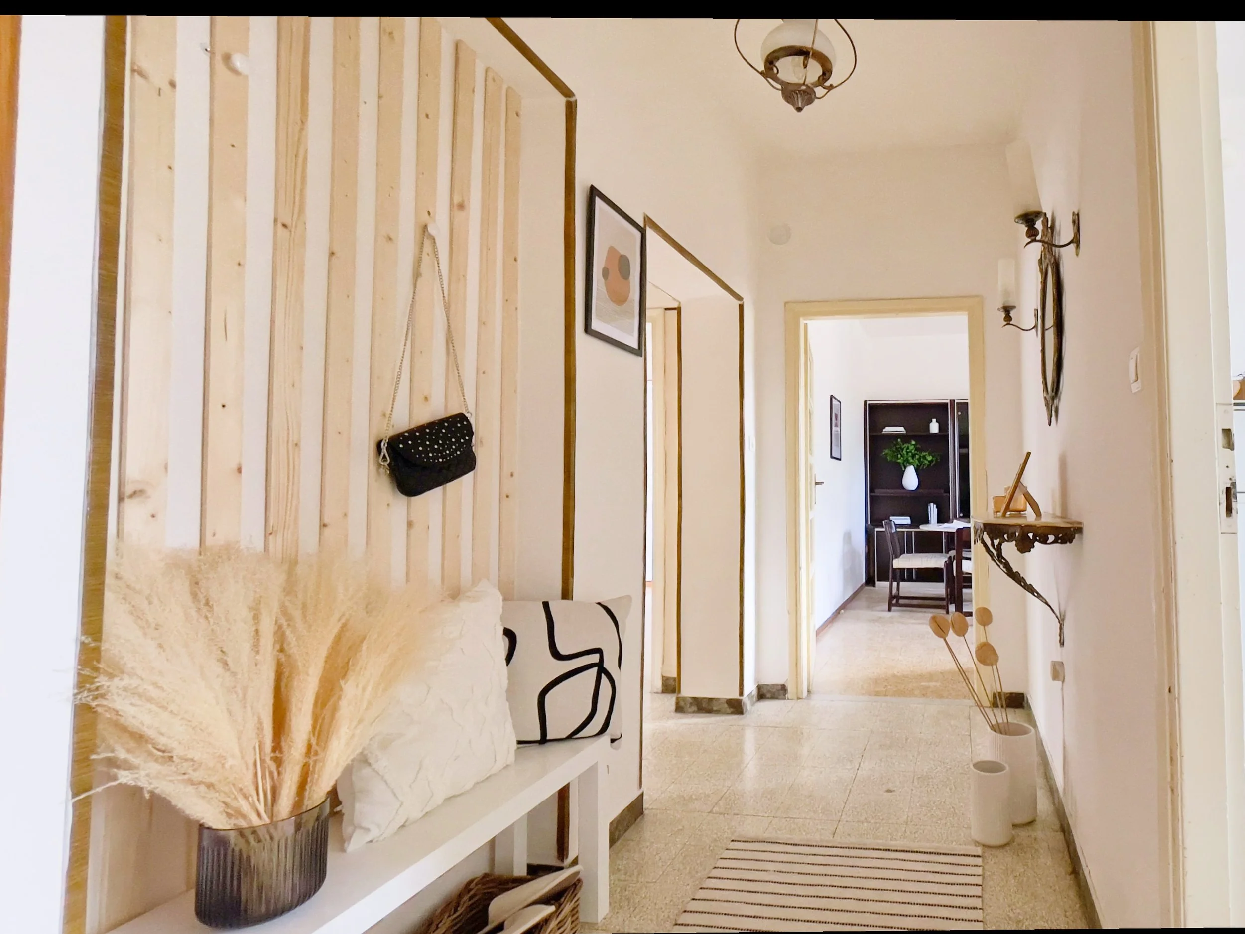 Corridoio interno di un appartamento con pareti bianche e un pannello decorativo in legno sulla sinistra. Ci sono elementi decorativi come una borsa appesa e un vaso con piante secche su una mensola. Verso il fondo, si intravede una sala con mobili e