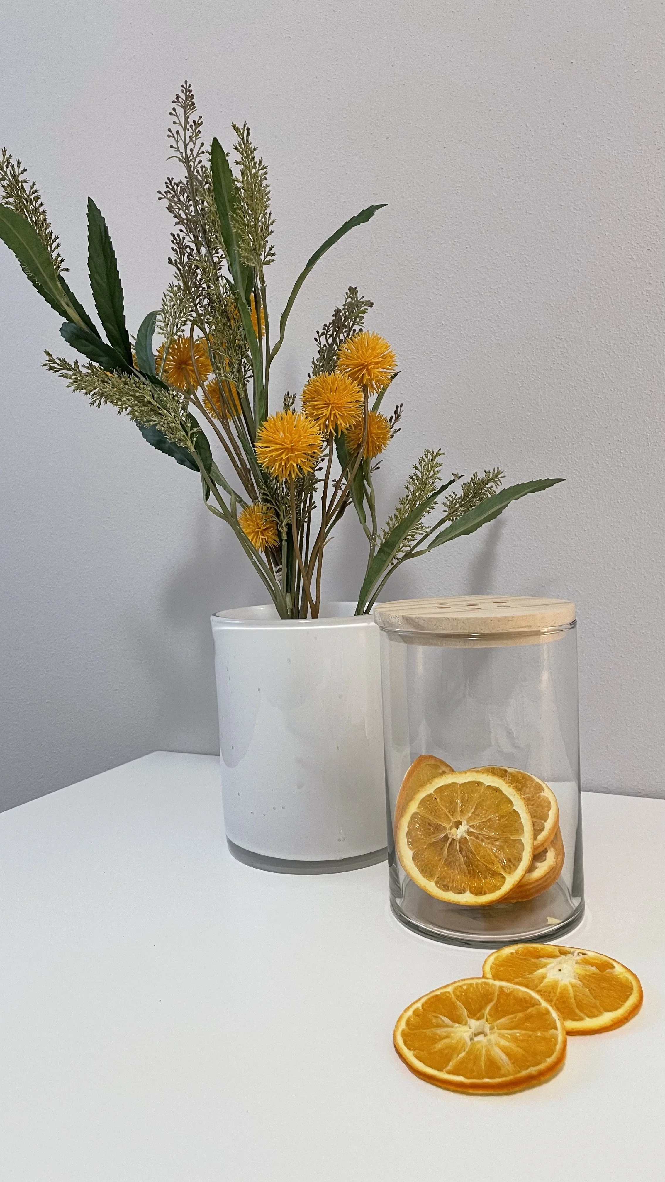 Vaso bianco con fiori gialli e barattolo con fette di arancia essiccate su un tavolo.