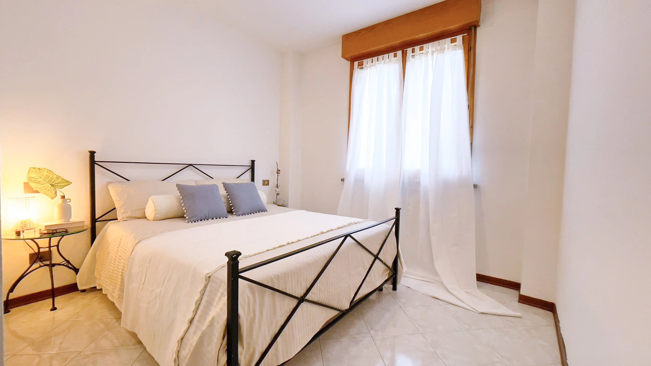 Camera da letto luminosa con letto matrimoniale, lenzuola bianche, cuscini grigi e tavolino con lampada e decorazioni. Finestra con tende bianche.