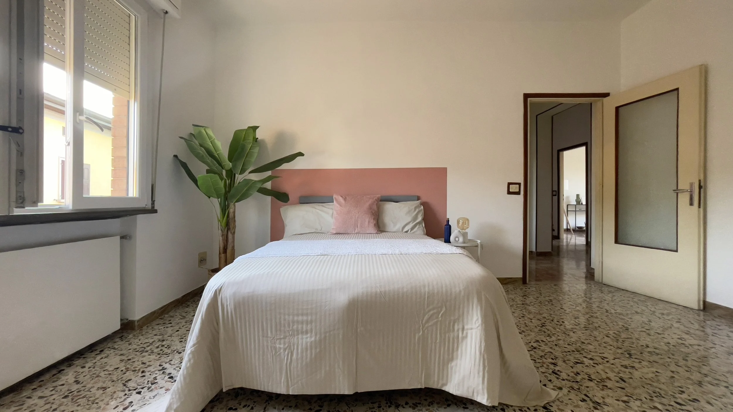 Camera da letto con letto matrimoniale, cuscino rosa, pianta verde a lato, finestra aperta a sinistra, e porta aperta a destra.