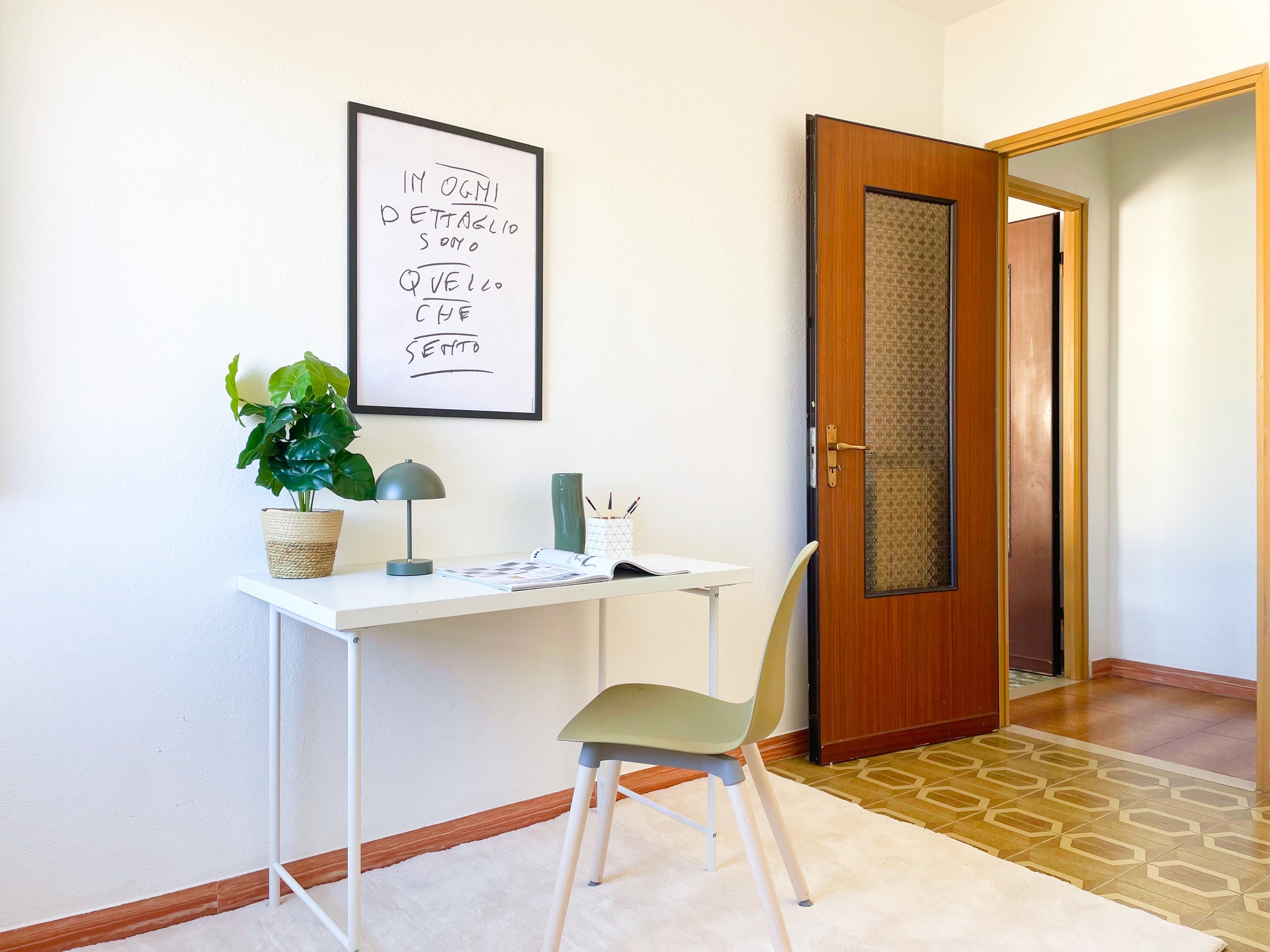Interno di una stanza luminosa con una scrivania bianca, una sedia verde chiaro, una pianta in vaso, una lampada da tavolo e un quadro con scritta appeso alla parete. La porta della stanza è aperta.